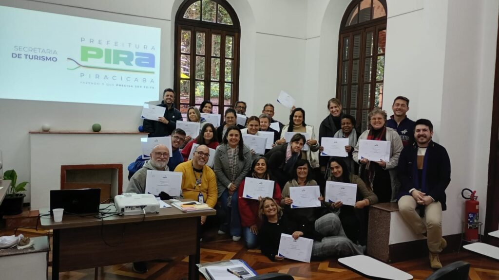 Nesta foto, um grupo de pessoas está reunido em uma sala, celebrando a conclusão de algum curso ou treinamento. Aqui estão os detalhes principais: A Celebração: Quase todas as pessoas estão segurando certificados de papel na mão. Elas parecem felizes e posam para uma foto em grupo. O Ambiente: É uma sala com piso de madeira, janelas altas com arcos e paredes brancas. À esquerda, há uma mesa com um projetor e um laptop. No fundo, um projetor exibe o logotipo da "Secretaria de Turismo - Prefeitura de Piracicaba". As Pessoas: Há um grupo grande e diverso, composto por homens e mulheres de várias idades, vestidos com roupas casuais e do dia a dia. Uma pessoa está sentada no chão, à frente do grupo, segurando seu certificado. Outros elementos: Há um extintor de incêndio vermelho no canto direito da sala. O clima geral é de realização e satisfação pelo encerramento de uma atividade.