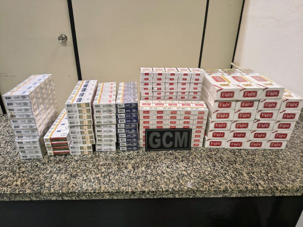 Esta imagem mostra uma grande quantidade de maços de cigarro organizados sobre um balcão de granito, com uma placa preta centralizada onde se lê "GCM" (Guarda Civil Municipal). Aqui estão os detalhes principais: Conteúdo: Diversas pilhas de maços de cigarro de marcas variadas, incluindo caixas maiores da marca "Eight" empilhadas à direita e maços menores (aparentemente da marca "Eight" e "Gift") dispostos em fileiras organizadas. Contexto: A presença da placa "GCM" sobre o balcão sugere que esta é uma apreensão realizada pela Guarda Civil Municipal. A organização dos itens indica que eles foram catalogados ou exibidos como prova de uma operação. Ambiente: O cenário parece ser uma delegacia, base operacional da guarda ou almoxarifado, com uma porta branca com maçaneta ao fundo. Disposição: Os maços estão meticulosamente alinhados, ocupando grande parte da superfície do balcão, destacando o volume total do material apreendido. Esta é uma imagem típica de registro de ocorrência policial envolvendo apreensão de mercadorias.