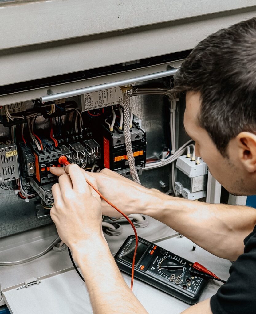 Nesta imagem, um técnico eletricista está concentrado realizando a manutenção de um painel elétrico. Ação principal: Ele utiliza uma ponta de prova vermelha de um multímetro digital para testar um dos contatores elétricos no interior do painel. O equipamento: O painel é composto por vários componentes eletrônicos, incluindo contatores (as peças cinzas e laranjas com terminais e fios) e uma fiação organizada. Há também um diagrama elétrico colado na parte superior do compartimento. O multímetro: Um multímetro digital preto está posicionado na base do painel, próximo às mãos do técnico, conectado aos cabos de medição. Detalhes: O técnico, que veste uma camiseta preta, está inclinado sobre o painel, demonstrando precisão no trabalho de diagnóstico ou verificação de voltagem/continuidade.