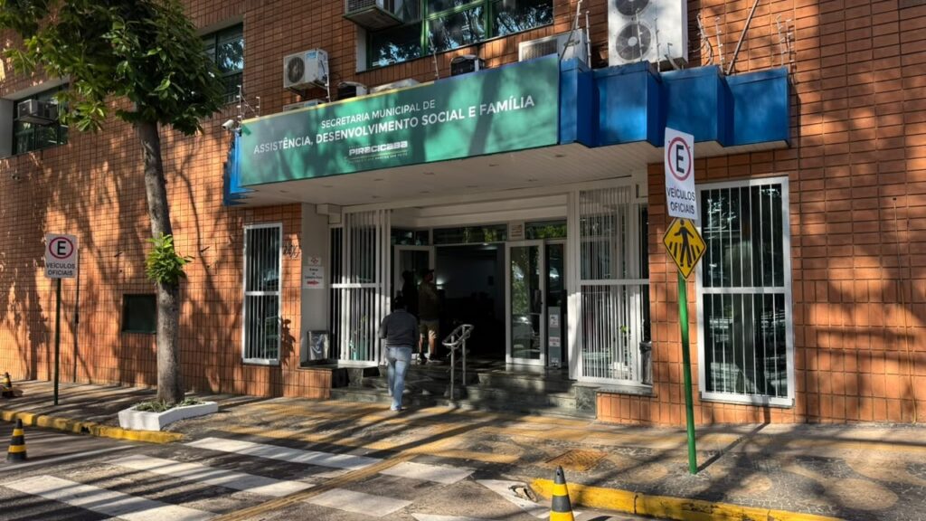 Fachada de prédio público vista de frente em dia ensolarado. Placa principal: faixa verde acima da entrada com o texto "SECRETARIA MUNICIPAL DE ASSISTÊNCIA, DESENVOLVIMENTO SOCIAL E FAMÍLIA" (nome da unidade municipal). Fachada e entrada: parede revestida com tijolos cerâmicos laranja; porta de vidro dupla com grades brancas laterais e pequenas escadas com corrimão de metal; também há uma rampa de acesso. Calçada e faixa de pedestres: faixa de pedestres ao nível da rua com sinalização tátil/amarela para acessibilidade; piso em padrão decorativo junto ao meio-fio. Sinalização viária: duas placas indicando proibição de estacionamento com o texto "VEÍCULOS OFICIAIS" próximas à entrada; placa de travessia de pedestres. Vegetação e sombra: árvore plantada na calçada à esquerda projetando sombras sobre a fachada e o passeio. Equipamentos: unidades de ar-condicionado instaladas acima da faixa verde; cone de sinalização no meio-fio. Pessoas: duas ou três pessoas entrando no prédio — vistas de costas, sem identificação. Iluminação e clima: luz natural forte, sombras longas de árvores, aparência de manhã ou tarde com sol baixo. Cena transmite um ambiente institucional acessível e em funcionamento.