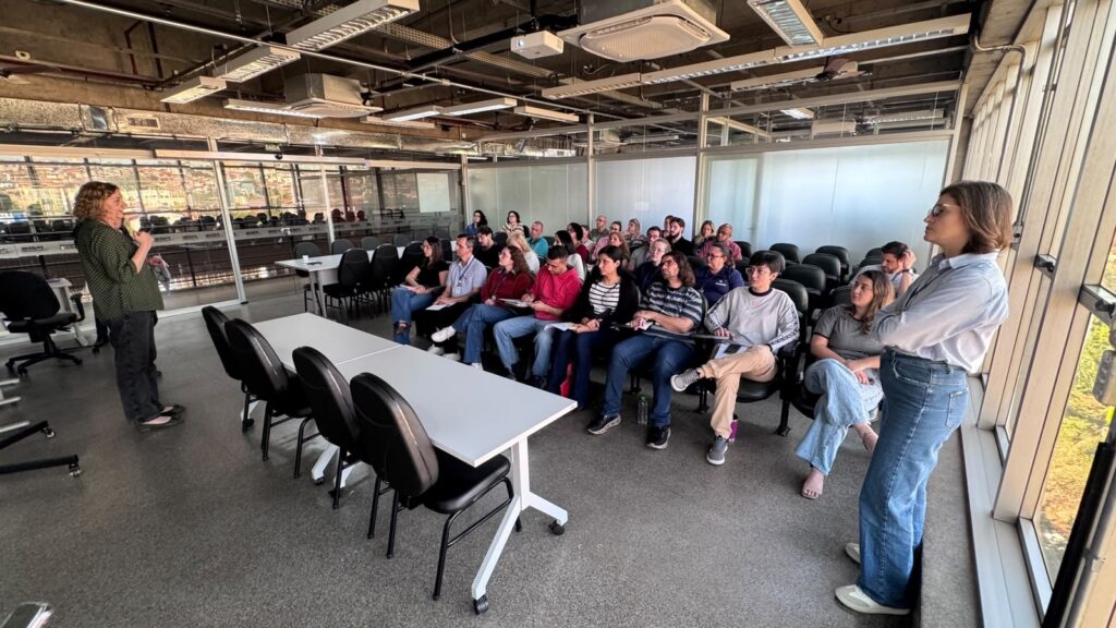 Foto de uma apresentação/treinamento em sala ampla e envidraçada. Cena geral: auditório pequeno ou sala de reunião com fileiras de cadeiras e mesas; grande janela à direita trazendo luz natural. Pessoas: cerca de 25–30 pessoas sentadas voltadas para a esquerda, prestando atenção; público misto (diferentes idades e gêneros). Duas mulheres em pé — uma à esquerda (provável palestrante) segurando microfone/objeto e falando para o grupo; outra à direita encostada no peitoril da janela, observando. Atitude e interação: plateia atenta, algumas pessoas fazendo anotações em cadernos; atmosfera parece formal e focada. Mobiliário e layout: cadeiras em fileiras com pequenas mesas dianteiras; duas mesas maiores e cadeiras vazias ao centro-esquerda; bancos acolchoados pretos. Piso liso e teto com fiação exposta e luminárias industriais. Iluminação e ambiente: iluminação mista — forte luz natural da janela direita e luz artificial do teto; reflexo das construções externas visível no vidro da parede traseira. Detalhes visuais: paredes de vidro separando a sala de outras áreas; itens pessoais como garrafa de água no chão junto a uma cadeira; roupas casuais-profissionais usadas pelos presentes (jeans, camisas).