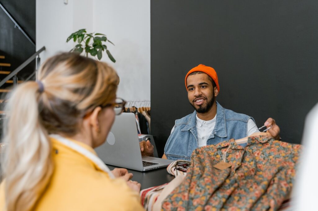 A imagem mostra uma cena em uma loja de roupas: um homem usando gorro laranja e colete jeans conversa com uma mulher de cabelo preso e casaco amarelo. Ele segura uma peça de roupa estampada em tons florais, enquanto um laptop está aberto sobre o balcão. O ambiente parece moderno e acolhedor, com ar de boutique.