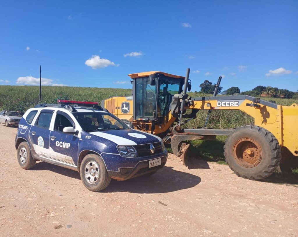 A foto mostra uma viatura da Guarda Civil Municipal está estacionada ao lado de uma motoniveladora recuperada em uma área rural, com plantação ao fundo.