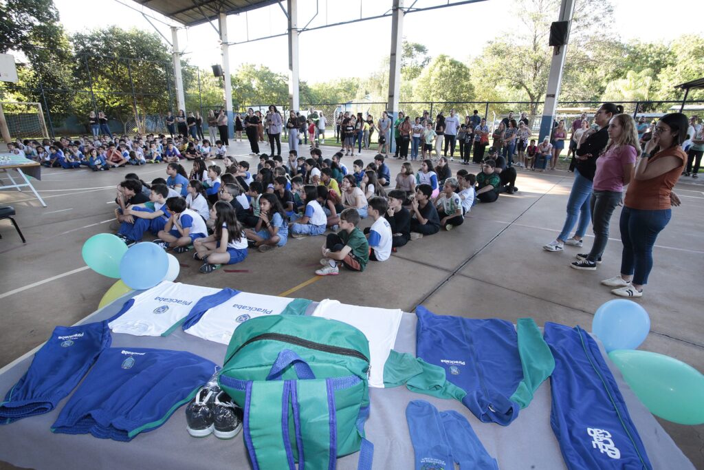 Foto de um evento escolar em uma quadra coberta, com alunos sentados no chão e público em pé ao fundo. Cena principal: várias crianças sentadas em filas no centro da quadra, olhando para a frente — parecem prestar atenção a uma apresentação. Primeiro plano: mesa com uniformes, mochilas e pares de tênis expostos; balões azuis e verdes e uma mochila verde em destaque sobre a mesa. Público adulto: pais, professores e outras pessoas em pé ao longo das laterais e ao fundo, observando a atividade. Ambiente: quadra poliesportiva coberta com estrutura metálica, grades nas laterais e árvores visíveis além da cerca; chão marcado com linhas de jogo. Iluminação: luz natural entrando pelas laterais, criando sombra suave; dia claro. Atmosfera: organizada e tranquila, típica de cerimônia escolar como entrega de materiais ou apresentação.