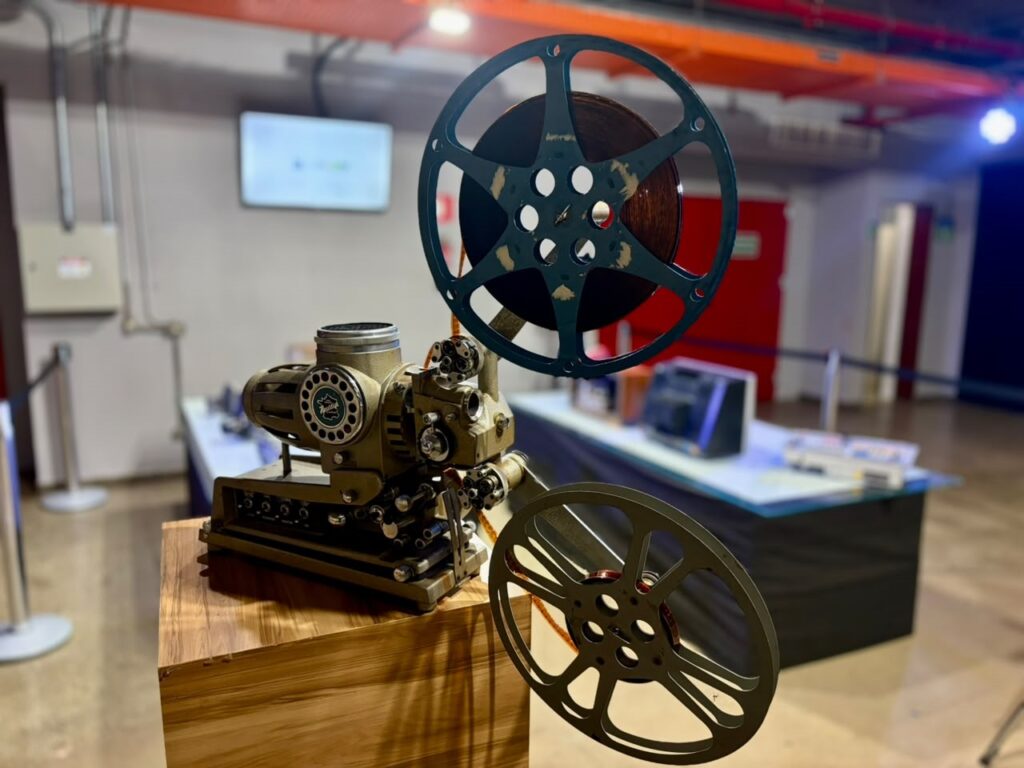 Esta é uma foto em destaque de um projetor de filmes clássico, montado sobre um pedestal de madeira com um acabamento amadeirado bonito. O Projetor: É uma peça central impressionante, com um design robusto e industrial, típico da era de ouro do cinema em casa ou em pequenas salas de projeção. Ele possui dois carretéis grandes (um na parte superior e outro na inferior) ainda com parte da película instalada. Detalhes: O corpo do aparelho é em um tom esverdeado/oliva, com engrenagens e mecanismos visíveis. Há um detalhe circular na lateral com um logotipo que parece ser da marca "Fujica". O Ambiente: A foto foi tirada em uma exposição cultural, pois, desfocado ao fundo, podemos ver outras mesas com mais equipamentos antigos (como a mesa de corte de vídeo que descrevemos anteriormente) e um ambiente amplo, sugerindo um museu ou galeria. É uma daquelas imagens que capturam perfeitamente a essência da tecnologia cinematográfica analógica!