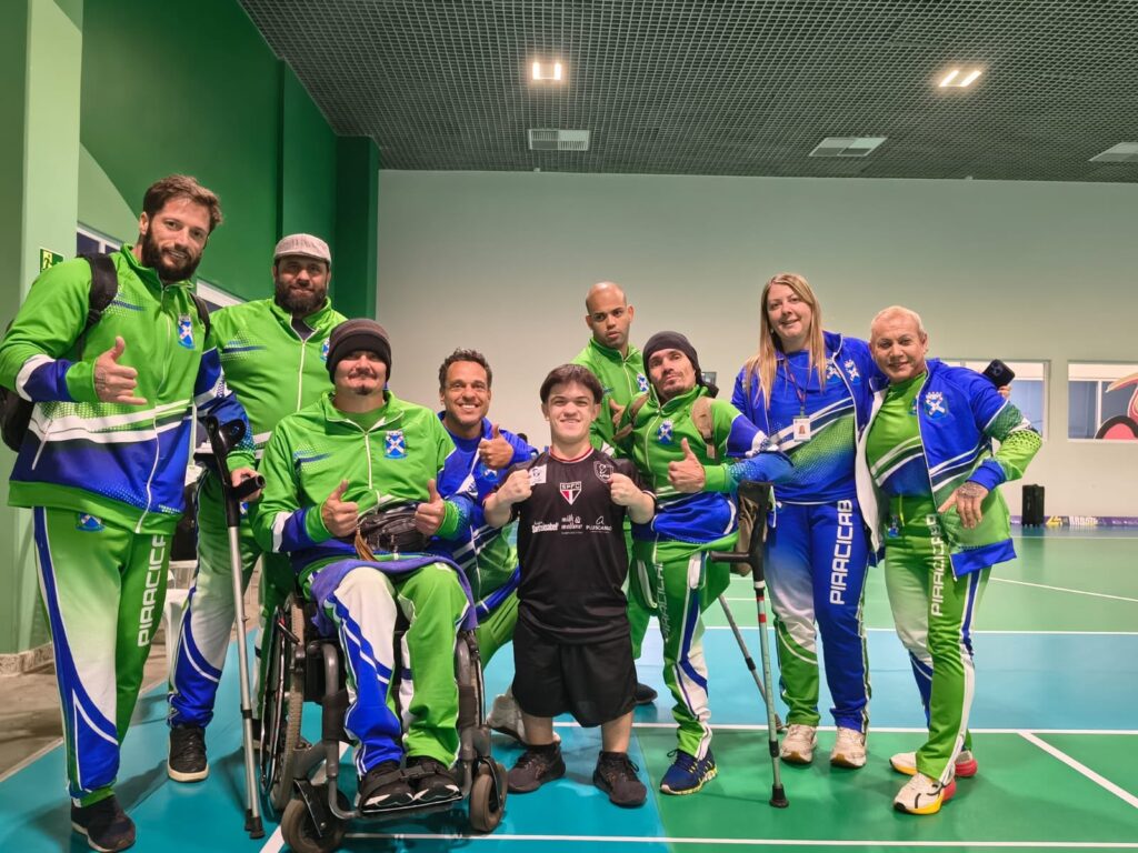 Grupo de dez pessoas posando em quadra esportiva coberta. Ambiente bem iluminado, piso e marcações típicas de ginásio/quadra esportiva. Pessoas e disposição Pessoas em dois níveis: maioria em pé formando semicírculo, duas pessoas em primeiro plano (uma em cadeira de rodas e outra agachada/ajoelhada no centro). No centro frontal, um jovem de camiseta preta (sorrindo) faz pose de vitória com os punhos cerrados. Diversos membros do grupo fazem gestos positivos (joinha, polegar para cima, sinal de hang loose). Roupas e identificação A maior parte usa uniformes esportivos verde e azul com a inscrição "PIRACICAB" na lateral da calça e um brasão no peito — indicam equipe ou delegação. Uma pessoa veste jaqueta azul predominante; o jovem central está com uniforme diferente (preto). Há equipamentos de mobilidade visíveis: cadeira de rodas esportiva, muletas e bengalas. Expressão e clima Atmosfera de celebração, união e camaradagem; expressões majoritariamente sorridentes e descontraídas. Linguagem corporal transmite orgulho e espírito de equipe, possivelmente após prova ou evento. Cenário e composição Fundo neutro com parede clara e teto técnico; o foco está no grupo centralizado sobre o piso da quadra. Foto bem enquadrada, boa nitidez e cores vivas (predomínio de verdes e azuis).