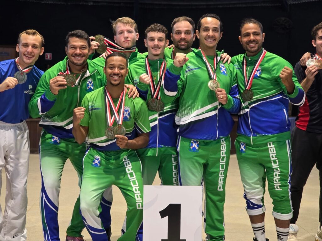 Nesta foto, vemos um grupo de atletas celebrando uma vitória esportiva, posando orgulhosos atrás de uma placa com o número "1". As Pessoas: São sete atletas masculinos principais no centro, todos usando medalhas no pescoço. Eles vestem uniformes coordenados em tons de verde, azul e branco, com a palavra "PIRACICABA" estampada nas calças e o brasão da cidade na camisa. As Ações: Eles estão sorrindo, alguns fazendo sinal de força com o braço ("joinha" ou punho fechado). O atleta que está no centro, à frente dos outros, segura duas medalhas. Detalhes: Há um atleta à esquerda que usa um uniforme azul, diferente dos outros, que parecem ser da equipe de São Carlos (dado o texto na camisa). O ambiente é uma quadra coberta. É uma imagem que transmite muita alegria e a sensação de conquista pelo primeiro lugar no pódio!