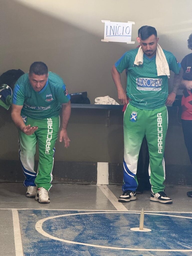 Esta é uma cena focada em um jogo de bocha ou malha, praticado em uma quadra interna. Os Jogadores: Dois homens estão em posição de jogo, ambos usando o mesmo uniforme esportivo: camisetas verdes com detalhes em azul e calças verdes com "PIRACICABA" escrito na perna. À esquerda, um jogador está agachado, concentrado, preparando-se para lançar (ou acabando de lançar) uma peça de metal. À direita, outro jogador está de pé, observando atentamente, com uma toalha branca pendurada no pescoço. O Ambiente: O chão é cinza com marcações brancas, e há um círculo azul pintado com um pino (bolim) no centro. Na parede atrás deles, há uma placa de papel afixada escrita "INÍCIO". Detalhes: O uniforme sugere que eles estão representando a equipe de Piracicaba em uma competição. A expressão dos jogadores é de total concentração.