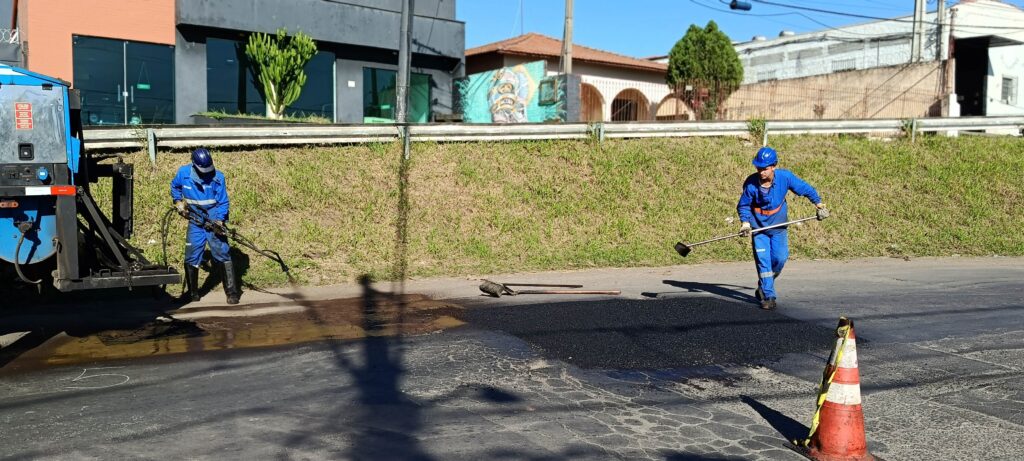 Dois trabalhadores vestindo macacões azuis e capacetes azuis estão consertando o asfalto de uma rua sob sol forte. Um deles, à esquerda, manipula uma mangueira ligada a um caminhão de obras — há uma poça escura de material fresco perto do veículo —; o outro, à direita, usa uma enxada ou régua longa para espalhar ou nivelar o novo recape. Há cones de sinalização laranja e fita de segurança no primeiro plano. Ao fundo há um pequeno talude gramado com uma mureta e, além dela, prédios comerciais e residenciais — em uma das paredes aparece um grafite colorido. A cena passa sensação de trabalho prático em rua urbana durante o dia, com sombras longas que sugerem manhã cedo ou final de tarde. O chão apresenta áreas remendadas e marcas de tráfego.