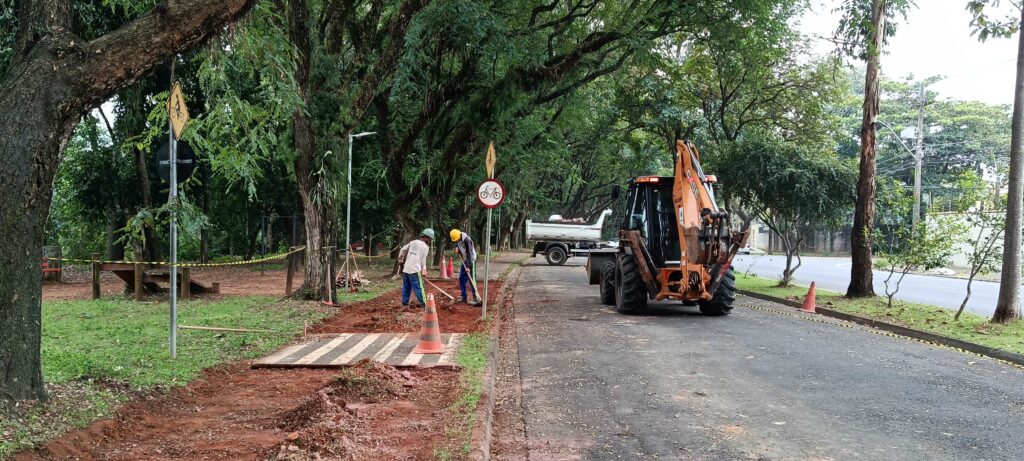 Esta é uma cena de trabalho de infraestrutura urbana, focada na construção ou reparo de uma calçada ou ciclovia. Ação: Dois trabalhadores, vestindo capacetes de segurança e roupas de trabalho, estão nivelando a terra vermelha na lateral da via. Eles utilizam ferramentas manuais (como enxadas ou ancinhos) para preparar o terreno. Equipamento: Há uma retroescavadeira grande (da marca Case) posicionada na pista, virada de costas para a câmera, pronta para auxiliar no serviço. Um caminhão basculante está parado logo à frente, possivelmente aguardando para retirar entulho ou carregar material. Sinalização: O local está sinalizado com cones de trânsito e uma placa de sinalização vertical de "trânsito de bicicletas" (com uma bicicleta desenhada dentro de um círculo vermelho), indicando que ali provavelmente é uma ciclovia ou uma área de compartilhamento. Ambiente: É um dia nublado em uma área urbana arborizada. Há muitas árvores grandes com troncos grossos à esquerda, criando uma área sombreada. O chão está sendo preparado para receber pavimentação, com uma parte de madeira temporária colocada sobre a terra para acesso. Detalhes: Há uma fita de sinalização amarela e preta delimitando a área de trabalho para segurança, e o asfalto da rua principal ao lado parece estar em boas condições. É um registro típico de manutenção viária, mostrando o esforço para melhorar a infraestrutura de pedestres e ciclistas na cidade.