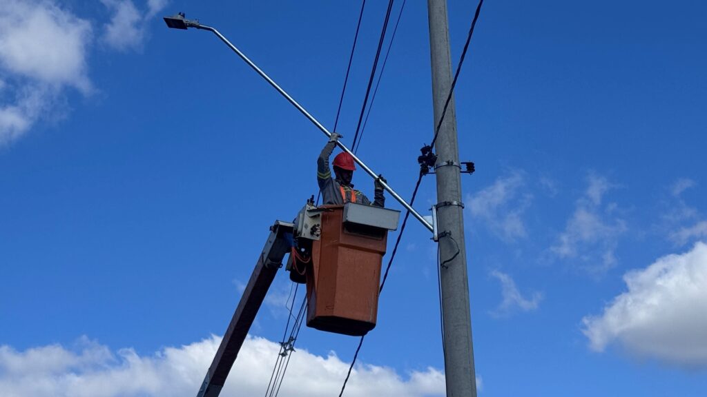 A imagem mostra um trabalhador realizando manutenção em poste de iluminação. Ele está dentro de uma plataforma elevatória laranja, usando equipamentos de segurança como capacete e colete refletivo. O profissional segura uma haste metálica próxima a um poste de energia, onde há fios e uma luminária instalada. O céu azul com algumas nuvens ao fundo indica um dia claro e tranquilo.