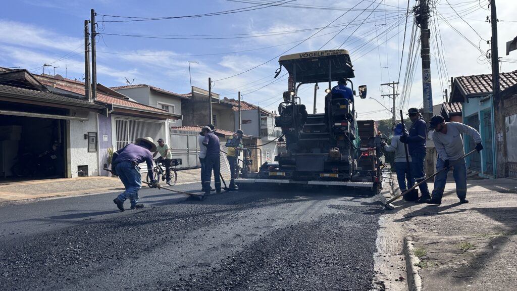 Equipe revitalizando o asfalto de uma rua residencial em dia claro. Ação principal: máquina pavimentadora (Vögele) no centro-direita despeja e compacta novo asfalto; vários operários ao redor espalham e nivelam a massa com ancinhos e talhadeiras. Pessoas: cerca de seis a oito trabalhadores usando roupas de trabalho (camisas compridas, alguns com bonés/chapéu), concentrados na operação; um ciclista parado observa a cena. Ferramentas e equipamentos: pavimentadora acoplada a um caminhão (fornecendo asfalto), ancinhos/rodo metálico, recipientes e outros utensílios de obra. Rua e pavimento: camada fresca de asfalto escuro sendo espalhada sobre o antigo piso; fragmentos soltos na frente e sulcos indicando trabalho recente. Ambiente: trecho residencial com casas térreas de fachada com portões, calçadas de concreto e muro; postes com muitos cabos elétricos ao longo da rua. Luz e clima: céu azul com poucas nuvens, luz forte e sombras definidas — parece manhã ou início de tarde ensolarada. Sensação geral: cena de manutenção urbana rotineira, organizada e em andamento, com foco na pavimentação e reparo da via.