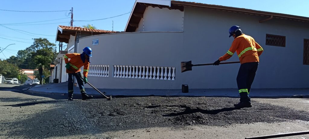 Nesta imagem, dois trabalhadores estão realizando um serviço de manutenção asfáltica em uma rua. Aqui estão os detalhes principais: Ação: Eles estão trabalhando com massa asfáltica, usando pás para espalhar e nivelar o material diretamente sobre o solo da via. Uniforme e Equipamento: Ambos vestem camisas de mangas longas na cor laranja fluorescente com faixas refletivas, calças escuras, luvas de proteção e capacetes de segurança azuis. Cenário: O trabalho ocorre em um dia ensolarado. Ao fundo, vê-se uma casa de cor clara com uma cerca de balustrada branca e uma placa de sinalização de rua indicando "Romário Nery". Há também um pequeno galão preto no chão próximo à parede da casa.