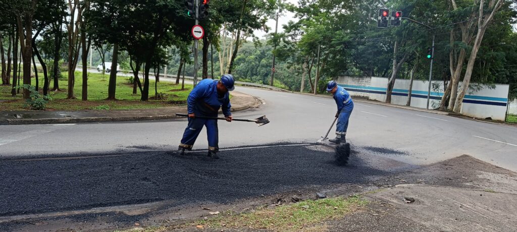 Foto de recapeamento de trecho de via urbana, com dois trabalhadores espalhando asfalto. Ação: dois operários em macacões azuis e capacetes trabalham com pás e enxadas para nivelar e espalhar uma camada de asfalto quente no centro da pista. Material/equipamento: mistura asfáltica preta recém-aplicada; não há máquina visível no primeiro plano — só as ferramentas manuais. Posição e enquadramento: tomada ampla e horizontal mostrando a faixa de asfalto no chão em primeiro plano e a rua curvando para a direita ao fundo. Sinalização e trânsito: sem tráfego próximo; semáforos visíveis ao fundo (um com luz vermelha e outro com verde) e placa de sentido obrigatório (seta para a direita) em um poste. Ambiente: área arborizada à esquerda com gramado e árvores; muro branco com faixa azul à direita; calçada e guia entre as árvores e a via. Clima/iluminação: dia nublado ou com luz difusa — cores amenas e sombra suave.