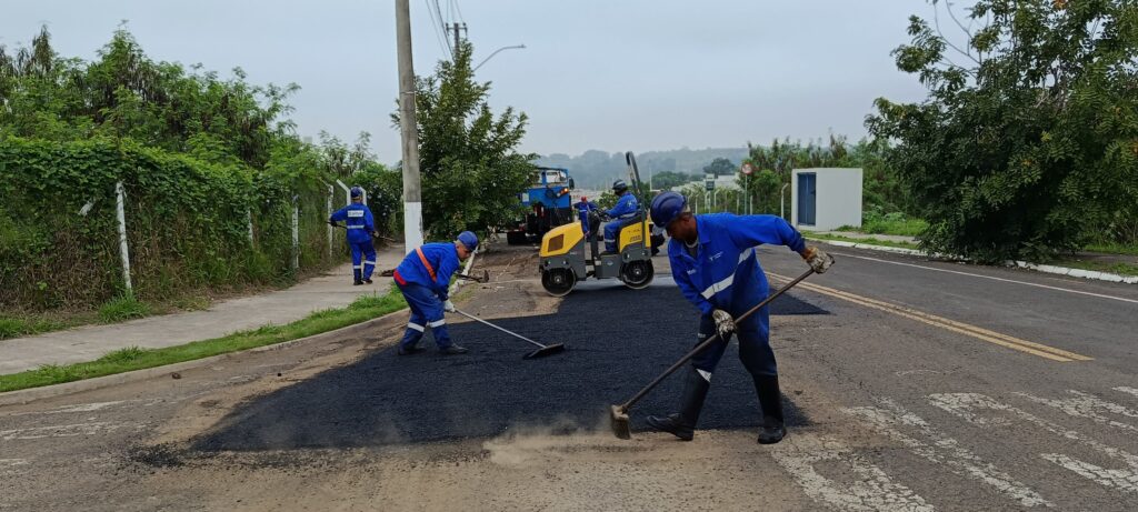 Esta é uma cena de trabalho de manutenção viária, onde uma equipe está realizando o recapeamento asfáltico de uma via. Aqui estão os detalhes: A Ação: Os trabalhadores, vestidos com uniformes azuis, capacetes e botas de segurança, estão espalhando e nivelando uma camada de asfalto novo sobre a pista. Um deles usa uma vassoura grande para distribuir o material, enquanto outro usa uma ferramenta de nivelamento. O Equipamento: Ao fundo, um operador conduz um rolo compactador amarelo, fundamental para assentar o asfalto e deixar a pista lisa e resistente após a aplicação. O Cenário: É um dia nublado em um ambiente externo. Há um poste de luz, vegetação nas laterais e uma calçada próxima. No fundo, é possível ver uma placa de trânsito de velocidade (50 km/h) e uma estrutura branca, indicando uma área urbana ou industrial. A Equipe: Além dos que estão nivelando, há um trabalhador caminhando na lateral da via, próximo à cerca, acompanhando o serviço. É um trabalho braçal intenso e essencial para manter as ruas seguras e conservadas para o tráfego de veículos.