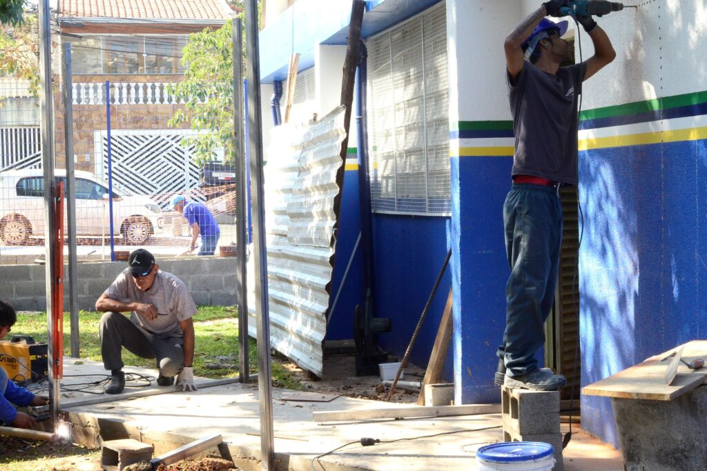 Obra ao ar livre vista de perto — três homens trabalhando na construção/recuperação de uma entrada ou calçada junto a um prédio pintado de branco e azul com faixas verde, branca, amarela e azul. Pontos principais: Pessoas: três operários em posições diferentes — um agachado no centro avaliando o piso (usa boné escuro, luva em uma mão), outro à direita em pé perfurando/instalando na parede (usa protetor auricular, capacete azul e luvas), e um terceiro mais ao fundo trabalhando no chão perto da grade. Ações: corte ou solda no canto inferior esquerdo (faíscas visíveis); medição/ajuste do contrapiso; perfuração na parede à direita. Materiais e ferramentas: blocos de concreto, perfurador/parafusadeira, máquina de solda/cortador, cabos elétricos no chão, régua/nível, balde de tinta/recipiente azul no primeiro plano. Estrutura: postes metálicos verticais e laje de concreto recém-colada; chapas onduladas provisórias apoiadas como proteção/isolamento entre o canteiro e a rua. Ambiente: fachada azul e branca de um edifício à direita; rua e casas ao fundo visíveis através da grade; gramado e sombra de árvores indicando dia ensolarado. Segurança: uso parcial de EPI (capacete, protetores auriculares, luvas), área delimitada por chapas e estrutura metálica; porém vários cabos e materiais no chão acrescentam risco de tropeço. Atmosfera: cena de trabalho prático e coordenado, em fase de instalação estrutural/acabamento externo.