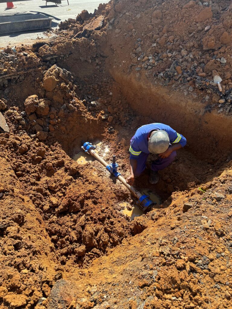 Escavação em via/terreno: vala rasa com terra argilosa avermelhada retirando parte do pavimento ao fundo. Trabalho em andamento em uma rede de tubulação enterrada. Pessoas e ação Um trabalhador agachado dentro da vala, de costas/parcialmente de perfil; veste uniforme azul com faixas reflexivas e capacete — não é possível identificar a pessoa. Ele manipula uma peça da tubulação próxima a um registro/união azul; parece ajustar ou verificar a conexão. Tubulação e equipamentos Tubo metálico/plástico com juntas e válvulas/uniões pintadas de azul visível na parte inferior da vala. Pequeno acúmulo de água lamacenta ao redor da conexão, indicando vazamento parcial ou água residual da escavação. Próximo à borda da vala há uma talocha/colher de pedreiro apoiada sobre o solo; bandejas e cones de sinalização no pavimento ao fundo. Solo e condições Solo argiloso, cor avermelhada, com pedras e fragmentos de pavimento misturados; bordas da vala mostram camadas de terra e entulho. Terreno seco na superfície, mas presença de lama e água no ponto de trabalho.