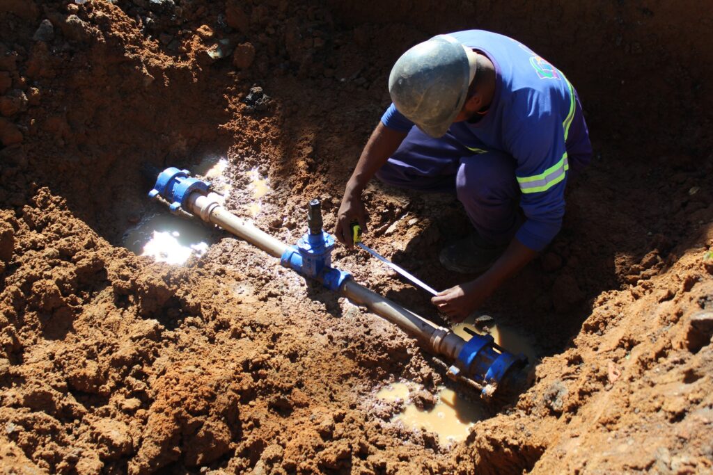Visão geral: homem trabalhando em uma vala rasa, consertando ou instalando uma tubulação de água. Foto em plano aberto mostrando solo argiloso vermelho ao redor e parte do encanamento exposto. Pessoa: trabalhador agachado de perfil, com capacete de proteção (sujo), roupa azul com faixas reflexivas nas mangas; rosto virado para baixo — não é possível identificar a pessoa. Tubulação e equipamentos: canos metálicos conectados por uniões e válvulas pintadas de azul; há um registro/torneira e um acoplamento rosqueado visível. O trabalhador segura uma fita métrica esticada ao longo do cano. Solo e condições: terra úmida e lamacenta no fundo da vala, com poças pequenas de água ao redor do trecho encanado; superfícies irregulares e cúmulos de terra escavada. Iluminação e clima: iluminação natural forte, sombras bem definidas — aparenta ser dia ensolarado. Contexto provável: manutenção hidráulica ou instalação de rede de água em área externa (obra pública ou obra civil), trabalho manual com medição e ajuste de conexões.