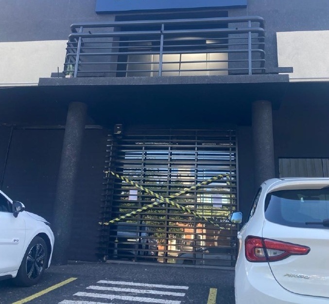 Esta imagem mostra a fachada de um estabelecimento comercial ou prédio de escritórios que parece estar temporariamente fechado ou em manutenção. Aqui estão os detalhes: Entrada: O acesso principal é bloqueado por uma grade metálica de correr, na qual foi colocada uma fita de isolamento amarela e preta em formato de "X". Fachada: O prédio possui uma estética moderna, com paredes pintadas em um tom escuro (cinza chumbo ou preto). Acima da entrada, há uma varanda ou sacada com grades metálicas. Carros: Dois veículos brancos estão estacionados, um de cada lado da entrada (apenas as laterais e traseiras são visíveis). O carro à esquerda exibe um emblema na lateral que parece ser da marca BYD. Piso: O chão indica um estacionamento ou área de acesso, com marcações de faixas amarelas e uma faixa de pedestres pintada. Avisos: Há pequenos papéis brancos fixados na grade metálica, provavelmente contendo comunicados ou informações sobre o fechamento do local. É uma cena comum de fachada urbana, sugerindo que o acesso ao interior está proibido ou restringido no momento da foto.