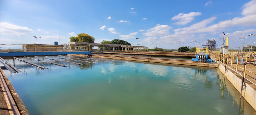 A imagem exibe uma vista ampla de uma estação de tratamento de água industrial sob um céu azul vibrante com nuvens esparsas. A cena principal é dominada por grandes tanques ou bacias de tratamento de água de formato retangular, preenchidos com água de tonalidades que variam do azul-celeste ao verde-azulado, especialmente mais azul distante e mais verde perto do primeiro plano. A água reflete claramente o céu azul com as nuvens brancas, criando um efeito espelhado, e também as estruturas circundantes. No tanque mais próximo, à esquerda, há várias fileiras de equipamentos horizontais, possivelmente barras de aeração ou de mistura, que se estendem pela superfície da água, criando pequenas ondulações e reflexos prateados onde a água se move. Ao longo das bordas dos tanques, estruturas robustas de concreto formam passarelas e plataformas de serviço. Estas passarelas são protegidas por corrimãos metálicos na cor amarela vibrante, que contrastam com o concreto bege-acinzentado e às vezes desgastado. À esquerda, uma ponte azul e amarela se estende sobre o tanque, apoiando uma série de tubulações horizontais que parecem ser parte do sistema de tratamento. No canto superior esquerdo, sobre uma plataforma, é visível um pequeno reservatório cilíndrico azul. No lado direito da imagem, uma complexa rede de tubulações, válvulas com volantes de controle e equipamentos azuis se estende ao longo da borda do tanque, indicando a natureza industrial da operação. Postes de iluminação de cor cinza-escura, equipados com múltiplas luminárias que se projetam em diferentes direções, estão distribuídos pelas plataformas e ao redor da instalação. Cabos elétricos pretos e outros conduítes atravessam o espaço, ligando diferentes partes da infraestrutura. Mais ao fundo, uma série de estruturas de concreto com colunas e telhados planos, talvez abrigando equipamentos ou escritórios, são visíveis. Atrás dessas estruturas, e ao longo do horizonte, há uma faixa de árvores com folhagem verdejante, quebrando a rigidez das construções industriais. O céu, que ocupa quase metade da imagem, é de um azul intenso com grandes nuvens brancas e fofas, sugerindo um dia ensolarado e claro. A luz do sol é forte, criando sombras nítidas e realçando as cores dos elementos. Não há pessoas ou animais visíveis na imagem. A atmosfera é de funcionalidade e tranquilidade, com a vastidão dos tanques e a calma da água contrastando com a complexidade da maquinaria.