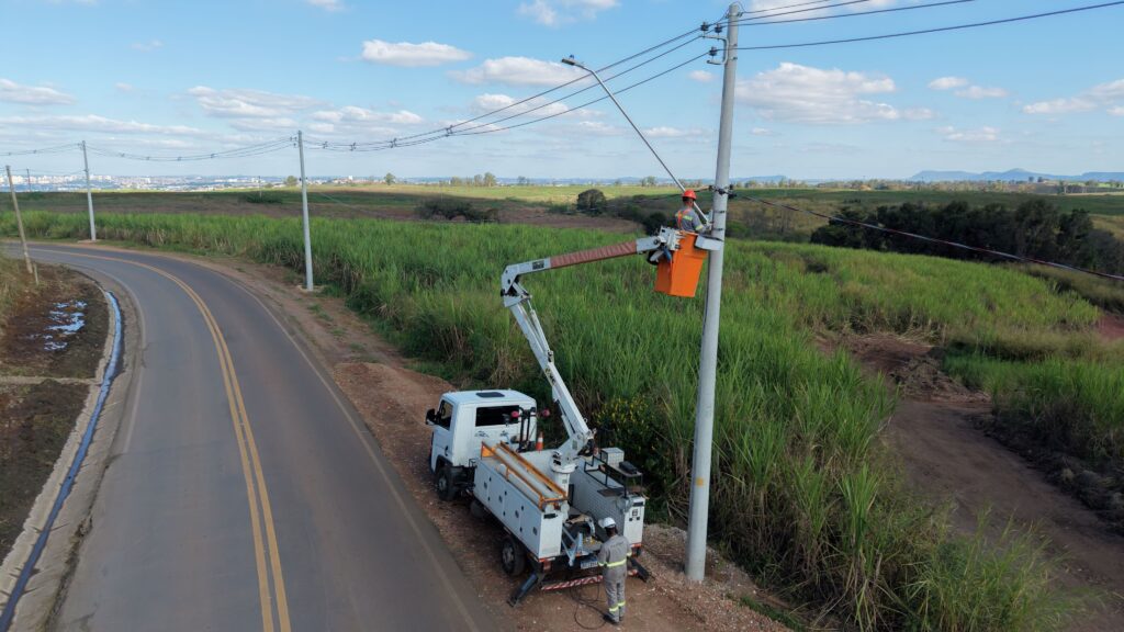 A imagem mostra uma equipe de manutenção elétrica trabalhando em uma área rural. Um caminhão com plataforma elevatória está estacionado ao lado de uma estrada asfaltada, e um trabalhador, usando equipamentos de segurança, está dentro do cesto laranja realizando ajustes em um poste de energia. Outro profissional, também com uniforme e capacete, opera o sistema no solo.
