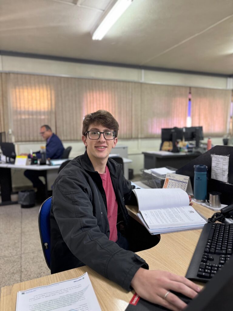 Um jovem sorridente está sentado em uma mesa de escritório com documentos e um computador. A imagem mostra um jovem com óculos e aparelho nos dentes, sorrindo para a câmera enquanto está sentado em uma cadeira de escritório azul, com o tronco virado para a direita. Ele veste uma jaqueta preta por cima de uma camiseta de cor rosa escuro. À sua frente, sobre uma mesa de madeira clara, está um livro aberto com textos e algumas folhas impressas, além de um teclado de computador preto e um telefone preto. No fundo, à direita, há uma garrafa térmica azul e um copo de metal. Um calendário também é visível sobre a mesa. Ao fundo, ao centro e à esquerda, vários cubículos de escritório são parcialmente visíveis, com mesas, computadores e persianas nas janelas, onde um homem mais velho está sentado em um cubículo à esquerda. As luzes do teto são visíveis no topo, proporcionando uma iluminação geral ao ambiente. A atmosfera é de um ambiente de trabalho rotineiro, mas o sorriso do jovem sugere um humor positivo e um momento de interação com quem tira a foto.