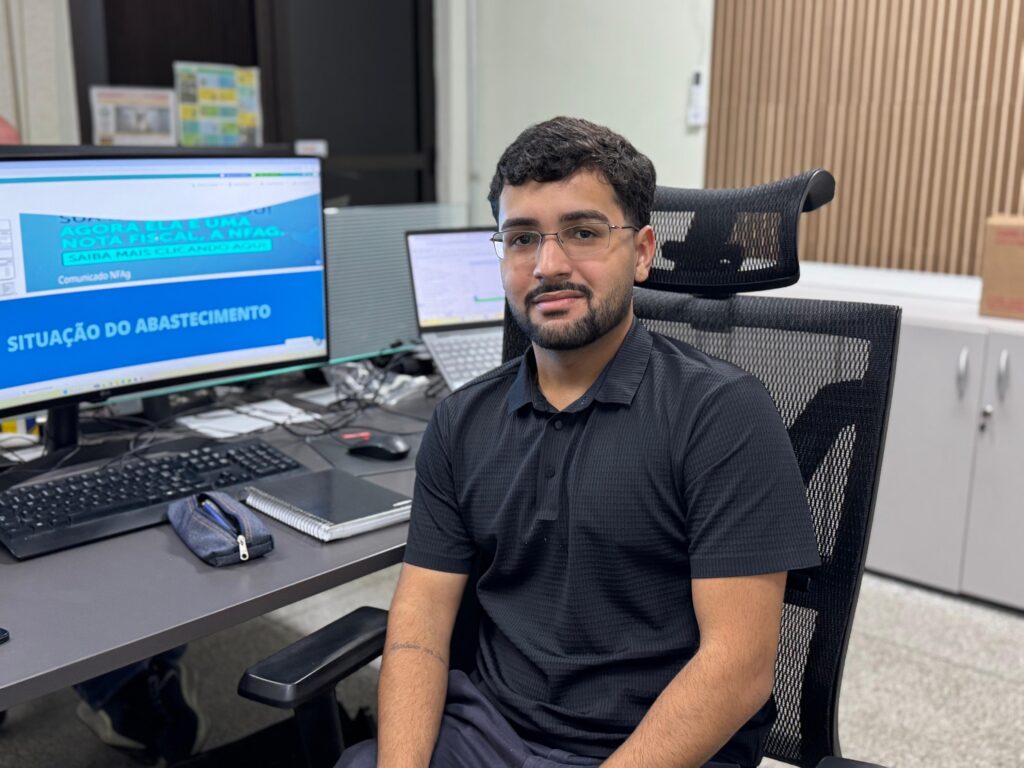 Um homem de óculos senta-se em uma cadeira de escritório em frente a um computador com informações de frete em sua tela. A imagem mostra um homem com barba e óculos, vestindo uma camisa polo preta de manga curta, sentado em uma cadeira de escritório preta com assento e encosto em malha. Ele está em um ambiente de escritório, com uma mesa à sua frente que contém vários objetos. Um monitor de computador grande exibe um texto azul em uma tela branca, com a maior parte do texto em português, incluindo "SITUAÇÃO DO ABASTECIMENTO" e "Comunicado NF Ag". Há também um teclado preto, um mouse vermelho e preto, cadernos e um estojo de lápis azul. Ao lado do monitor principal, há outro monitor e um laptop. A cadeira de escritório está posicionada de forma que o espectador tem uma visão clara do homem e de sua estação de trabalho. O fundo da sala de escritório apresenta uma parede com painéis verticais de madeira bege e um gabinete branco com alças prateadas em um canto. A iluminação parece ser artificial, típica de um ambiente interno de escritório. O humor geral da imagem é profissional e direto, capturando um momento de trabalho em um escritório. Vindo de um close mais próximo, uma tatuagem leve é visível no antebraço do homem.