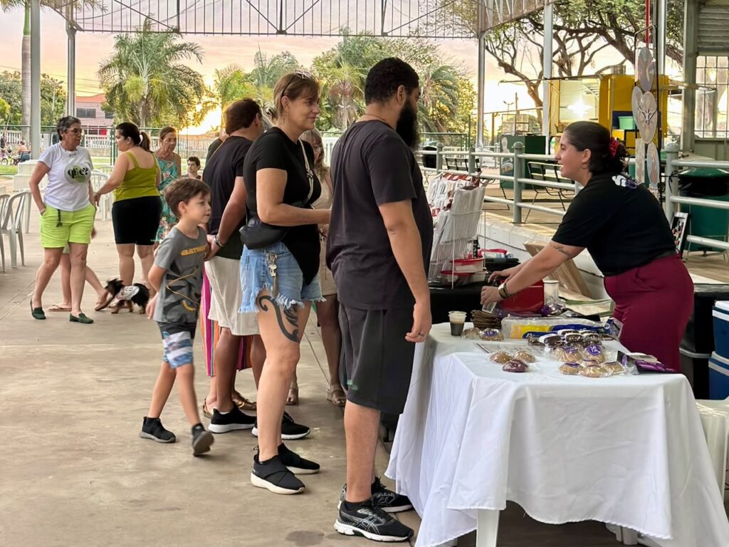 Esta imagem mostra um momento de interação em uma feira ou evento ao ar livre, possivelmente em um pavilhão coberto. A cena principal: À direita, uma mulher com uma camiseta preta atende um casal em uma mesa coberta por uma toalha branca. Na mesa, há vários produtos embalados, que parecem ser doces ou biscoitos artesanais. As pessoas: O casal está de costas ou de lado: a mulher usa camiseta preta e shorts jeans, e o homem veste uma camiseta cinza escura e bermuda preta. Um menino pequeno, com uma camiseta estampada do Batman, observa a cena ao lado deles. Ao fundo, à esquerda, outras pessoas caminham pelo local. Vemos uma mulher de camiseta branca e shorts neon verde, outra de regata verde e, inclusive, um cachorrinho pequeno caminhando junto ao grupo. Ambiente: O espaço é amplo e parece ser um local público. A luz sugere final de tarde, com um tom alaranjado no céu ao fundo.