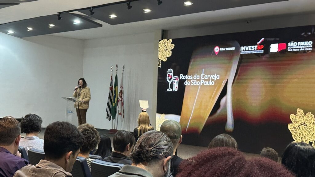 Foto de um evento em auditório com foco numa palestra ou anúncio. Cena geral: público sentado de costas na parte inferior da imagem, assistindo a um palco à esquerda. O auditório tem teto com spots de luz embutidos e paredes claras. Palco: à esquerda, uma mulher em pé atrás de um púlpito de acrílico, falando ao microfone. Ela veste casaco bege/mostarda e calça em tom escuro. Não é possível identificar quem é. Elementos no palco: ao lado do púlpito há três mastros com bandeiras (entre elas a bandeira do Brasil e bandeiras do estado/município). O púlpito tem um logotipo pequeno visível na base. Telão/painel: à direita ocupa grande parte da imagem — tela com a arte do evento onde se lê “Rotas da Cerveja de São Paulo”. Também aparecem os logos “INVESTSP”, “SP PRA TODOS” e o brasão/identidade visual do Governo de São Paulo. A imagem do fundo mostra, em grande plano, um copo de cerveja e elementos decorativos em dourado. Público: várias pessoas de idades variáveis, sentadas em filas, algumas com cabelos grisalhos; ninguém está em destaque ou identificável. Tom/atmosfera: profissional e formal, parece uma apresentação institucional ou lançamento relacionado ao turismo/mercado cervejeiro em São Paulo. Iluminação neutra, ambiente interno e atenção do público voltada para a palestrante e o painel.