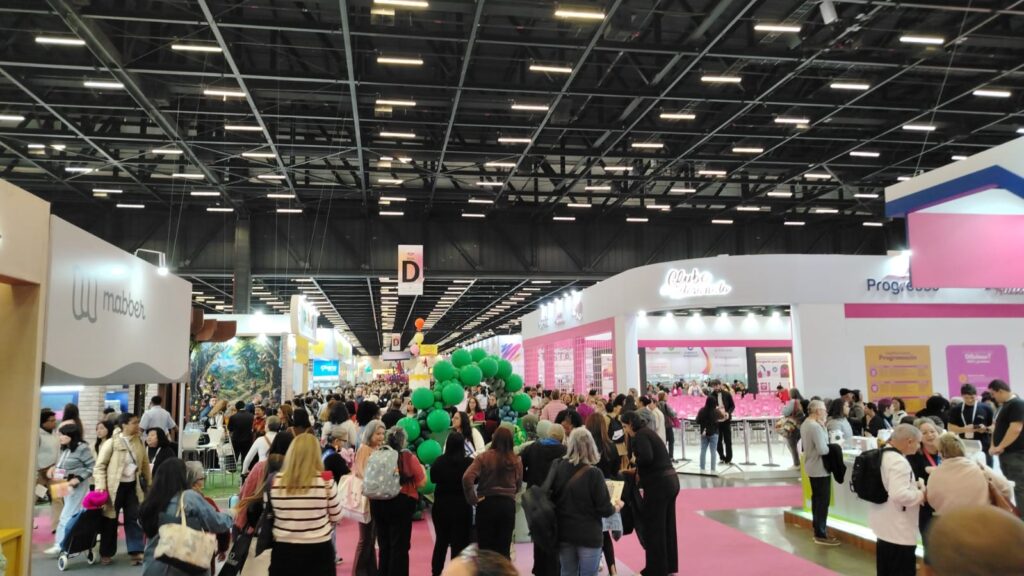 A imagem mostra um grande pavilhão de exposições durante um evento ou feira comercial bastante movimentada. Há muitos visitantes caminhando pelos corredores largos, conversando e visitando os estandes. Alguns detalhes que se destacam: Ambiente: Um centro de convenções amplo, com pé-direito alto, estrutura metálica aparente no teto e iluminação distribuída por todo o espaço. Movimento: O corredor principal está cheio de pessoas de diferentes idades, muitas usando crachás, carregando bolsas, mochilas e sacolas, o que sugere um evento profissional ou especializado. Estandes: Nas laterais há diversos estandes de empresas com identidade visual própria. Sinalização: No centro superior da imagem há uma placa indicando o setor "D", facilitando a orientação dos visitantes. Decoração: Próximo ao centro do corredor há um arco formado por balões verdes, que parece marcar uma atração, entrada ou ponto de destaque do evento. Cores: O piso possui faixas de carpete rosa que delimitam os corredores e contrastam com os tons neutros do ambiente. No conjunto, a fotografia transmite a impressão de um evento de grande porte, bem organizado e com alta circulação de público, provavelmente uma feira de negócios, artesanato, empreendedorismo ou produtos especializados.
