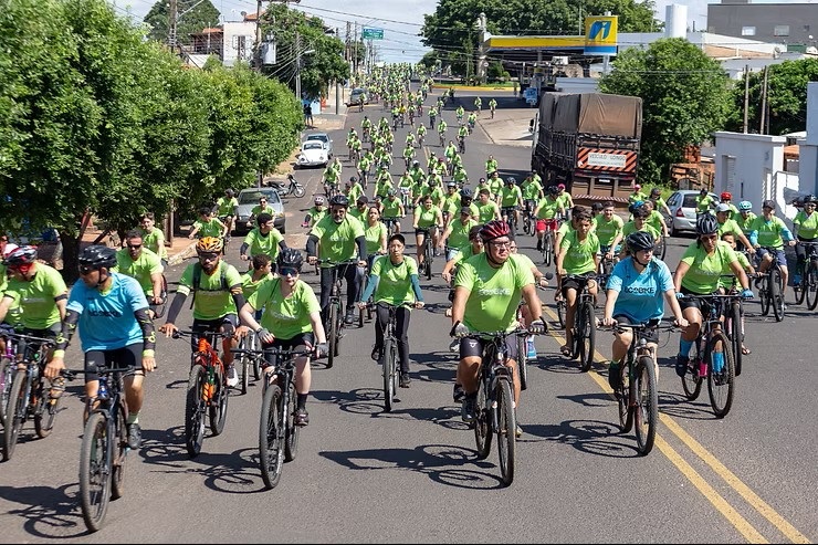 Foto de um grande grupo de ciclistas participando de um passeio organizado em via urbana. Observações: Centenas de pessoas em bicicletas, distribuídas pela largura da rua e estendendo-se ao fundo da imagem. A maioria veste camisetas verdes fluorescentes; alguns ciclistas usam camisetas azuis. Muitos usam capacetes e alguns óculos de proteção; é visível equipamento variado (mochilas, garrafinhas, bolsas de quadro). Bicicletas são em sua maioria mountain bikes/trekking, com pneus grossos e guidões retos. Rua asfaltada com pintura de faixa dupla amarela ao centro; sombras curtas indicam sol forte e horário próximo ao meio-dia. Ao fundo há um posto de combustível e alguns veículos estacionados/ em tráfego (um caminhão grande à direita). Laterais com arborização e construções residenciais/comerciais; ambiente parece organizado, sem sinais óbvios de tumulto. Atmosfera geral: evento coletivo, recreativo ou de conscientização, com grande participação e caráter seguro (uso de capacetes e camisetas padronizadas).