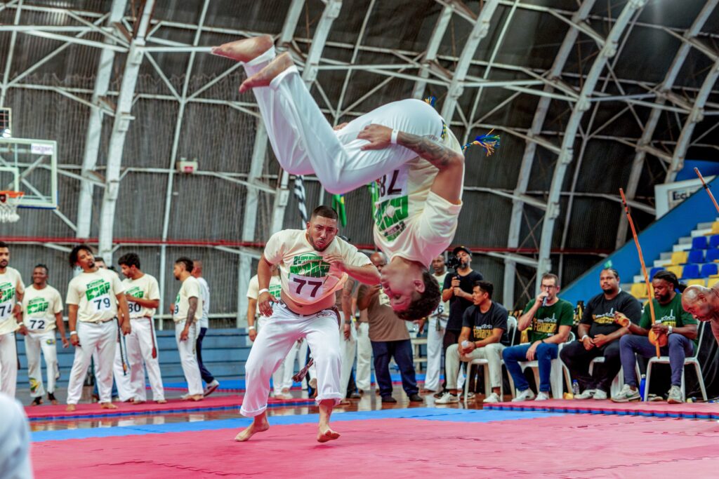 Esta é uma cena incrível de um evento de capoeira dentro de um ginásio! O Movimento: O destaque total é um capoeirista que está realizando um salto acrobático impressionante (como um mortal ou "au" sem mãos) no ar. Ele está de cabeça para baixo e com as pernas abertas, demonstrando muita habilidade e força. O "Roda": Logo abaixo, outro capoeirista, vestindo uma camiseta bege com o número "77" e calças brancas, parece estar no meio de um movimento de esquiva ou ginga, mantendo a atenção no jogo. Os Participantes: Ao fundo e nas laterais, há vários outros praticantes de capoeira vestindo uniformes padronizados (camisetas brancas ou beges com números, calças brancas), observando o movimento com foco. O Ambiente: O evento acontece em um ginásio coberto com piso de placas de EVA (azul e vermelho). À direita, sentados em cadeiras, estão os músicos, com berimbaus e outros instrumentos, garantindo a trilha sonora e o ritmo da roda. Há também algumas pessoas assistindo e um homem com uma câmera filmando a performance. Clima: A atmosfera é de competição e demonstração cultural, com muita energia e concentração de todos os envolvidos. É uma foto que captura perfeitamente a agilidade e a tradição da capoeira!