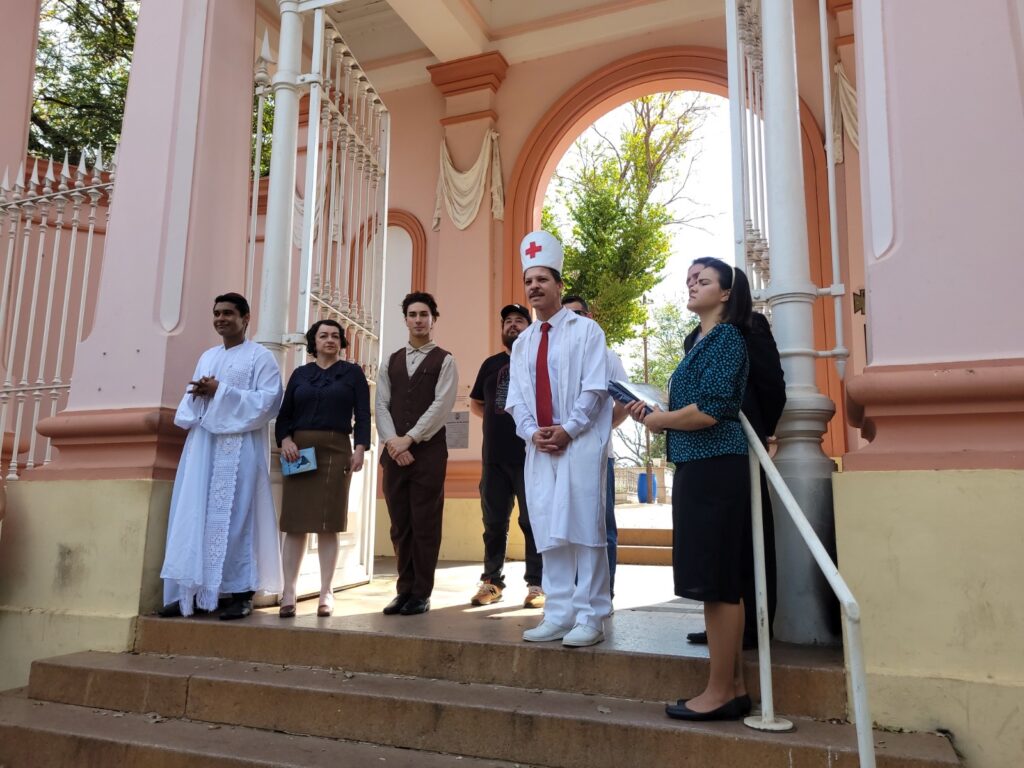 Foto de um grupo de pessoas posando/participando de uma cerimônia ou apresentação na entrada de um prédio com fachada cor de rosa (provavelmente uma igreja ou edifício histórico). Observações principais: Local: grande portal/entrada com arco e grades brancas; paredes em tons de rosa e detalhes arquitetônicos clássicos. Degraus de pedra levam ao patamar onde o grupo está em pé. Pessoas: sete ou oito pessoas alinhadas no patamar, em posições ligeiramente espaçadas, olhando em direções próximas (alguns para frente, outros levemente de lado). Trajes variados que sugerem personagens ou funções: À direita, um homem vestido de branco com jaleco e chapéu com cruz vermelha, gravata vermelha — aparência de enfermeiro/médico cerimonioso (fantasia ou traje simbólico). À esquerda, um homem com túnica branca longa, que lembra vestimenta religiosa ou litúrgica. Outras pessoas usam roupas formais ou figurinos: uma mulher de saia e blusa escura, um homem jovem com colete marrom estilo antigo, uma mulher com vestido/saia preta e blazer azul estampado segurando papéis ou livro. Ao fundo há um homem com roupa casual e boné, parcialmente visível. Ações/objetos: a mulher à direita segura pastas ou papéis — pode estar lendo ou organizando a cerimônia. Outra pessoa segura um pequeno livro/folheto. Não há gestos bruscos; postura majoritariamente séria ou atenta. Iluminação e clima: dia claro, luz natural; sombras indicam sol forte vindo de lado/trás. Cores quentes dominam pela arquitetura rosa e pelo tom da luz. Atmosfera: parece evento comunitário, encenação teatral, reconstituição histórica ou cerimônia simbólica — mistura de elementos religiosos e profissionais/figurinos que sugerem papéis distintos. Cena composta, ordenada e sem aglomeração ao redor.