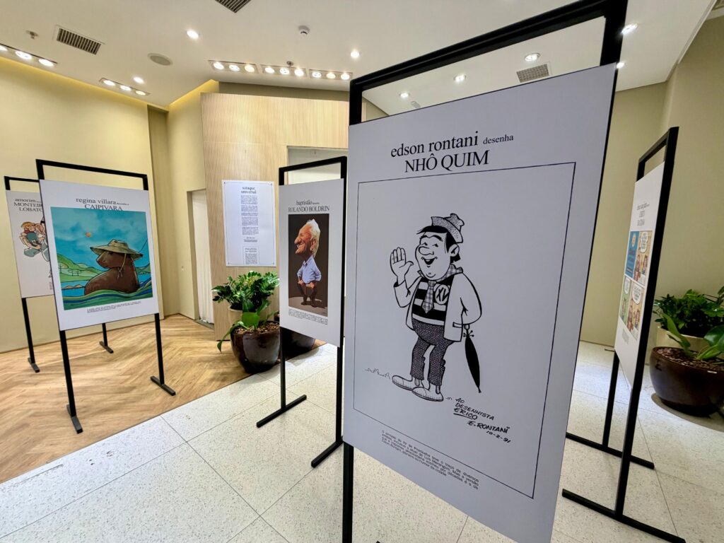 Esta é uma foto de uma exposição de arte em um ambiente bem iluminado e moderno, com foco em caricaturas e charges. Destaque principal: Em primeiro plano, há um painel vertical com uma caricatura em preto e branco do personagem "Nhô Quim", desenhada por Edson Rontani (com a data 10-2-91). O personagem está acenando, vestindo um chapéu, jaqueta e segurando um guarda-chuva. Outros painéis: À esquerda, há outros expositores. Um deles apresenta uma ilustração colorida de uma capivara de chapéu em uma paisagem rural, intitulada "Regina Villara desenha a Caipivara". Há também outro painel mostrando uma caricatura de Rolando Boldrin, feita por Baptistão. Ambiente: O local possui piso de pedra clara, paredes com detalhes em madeira ripada e plantas decorativas em vasos, criando uma atmosfera organizada e cultural. Há um texto informativo na parede ao fundo sobre "Sotaque Universal". A exposição parece celebrar o humor gráfico e a cultura local, possivelmente relacionada a Piracicaba, dado o personagem Nhô Quim.