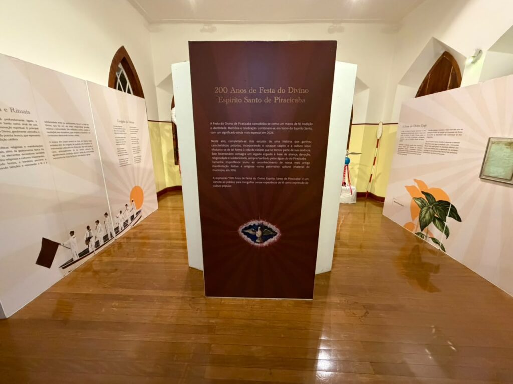 A imagem mostra uma exposição dentro de um espaço cultural, sobre a Festa do Divino Espírito Santo de Piracicaba.Detalhes da cena:Tema central: No meio da sala tem um totem marrom escuro com o título "200 Anos de Festa do Divino Espírito Santo de Piracicaba". O texto fala que em 2026 a festa completa 200 anos e que é um marco de fé, tradição e identidade da cidade. Menciona também que a festa mistura cultura caipira, religiosidade, solidariedade e patrimônio imaterial do município, reconhecido em 2016. Na base do totem tem a imagem da pomba branca, símbolo do Divino.Painéis: Ao redor tem painéis brancos com textos e ilustrações. À esquerda: ilustração de pessoas de branco em um barco com bandeira, e títulos como "Cortejo do Divino" e "Rituais".À direita: ilustração de folhas verdes e laranjas, com o título "A Festa do Divino Hoje". Os painéis usam tons de laranja, branco e raios de sol, passando leveza.Espaço: O local tem piso de madeira, paredes brancas e janelas em arco de madeira, típicas de prédios históricos. A iluminação é clara e o ambiente está organizado para visitação.Clima: A exposição celebra a história e a importância cultural e religiosa da Festa do Divino em Piracicaba, convidando o público a conhecer essa tradição de 2 séculos.