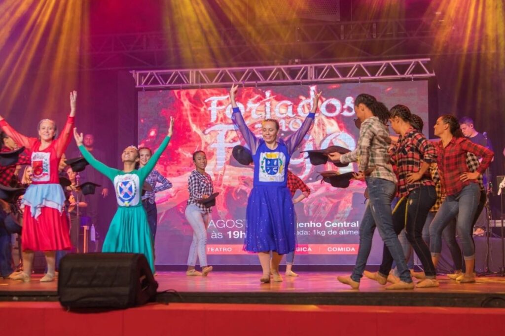 Esta é uma foto vibrante de uma apresentação de dança no palco de um teatro. Aqui estão os detalhes principais: O Grupo de Dança: Vários dançarinos estão no palco, muitos deles usando roupas que remetem a trajes típicos de festas caipiras ou quadrilhas, como camisas xadrez e calças jeans. Destaques: Algumas das dançarinas principais usam vestidos coloridos e temáticos, em tons de vermelho, azul e verde, com um emblema ou brasão estampado no peito, o que sugere uma apresentação cultural ou oficial. Elas estão com os braços erguidos em pose de dança. O Cenário: Ao fundo, há um grande telão LED exibindo uma imagem com tons de vermelho e fogo, com um texto que parece ser o título do evento (parte dele visível como "Feriados..." ou algo semelhante) e informações sobre data, local (Engenho Central) e entrada (1kg de alimento). Iluminação e Clima: A iluminação é dramática, com focos de luz amarela descendo do topo do palco, criando feixes de luz que dão destaque aos artistas. O ambiente parece ser de muita animação, movimento e celebração cultural. É uma cena que transmite muita energia e dedicação artística!