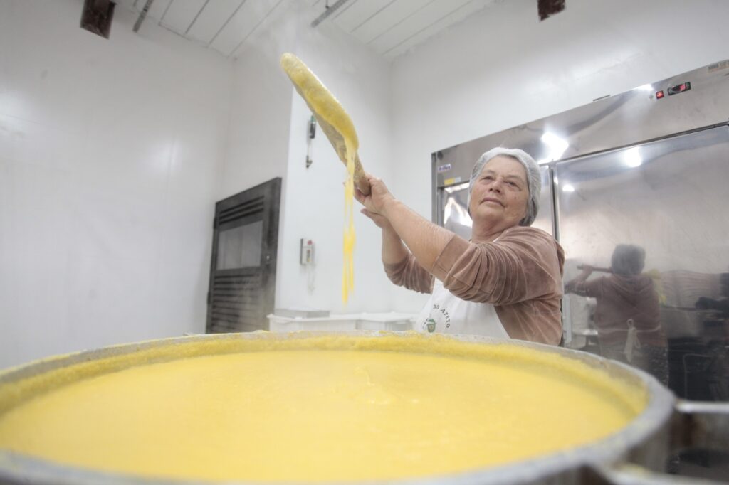 Foto de uma pessoa adulta preparando uma grande panela de polenta ou mingau amarelo em cozinha industrial. Observações principais: Pessoa de idade adulta/idosa usando touca descartável e avental branco, segurando uma pá/colher longa e levantando uma porção do alimento que escorre em fios. Grande panela metálica em primeiro plano, cheia de uma mistura cremosa e amarela (aparenta ser polenta, curau ou creme de milho), com vapor leve subindo. Ambiente limpo e profissional: paredes brancas, forro com ripas, e grande equipamento inox (possivelmente câmara fria ou forno) ao fundo. Iluminação forte e difusa, sem sombras duras; reflexos no aço inoxidável. Expressão concentrada e serena na face da pessoa; gesto indica experiência e técnica no manuseio do alimento. Detalhes de segurança/higiene visíveis: touca, avental; não há outros trabalhadores próximos na imagem. Composição: plano baixo com foco na superfície da panela em primeiro plano e na pessoa em plano médio-direito, transmitindo sensação de escala da panela e do trabalho artesanal.