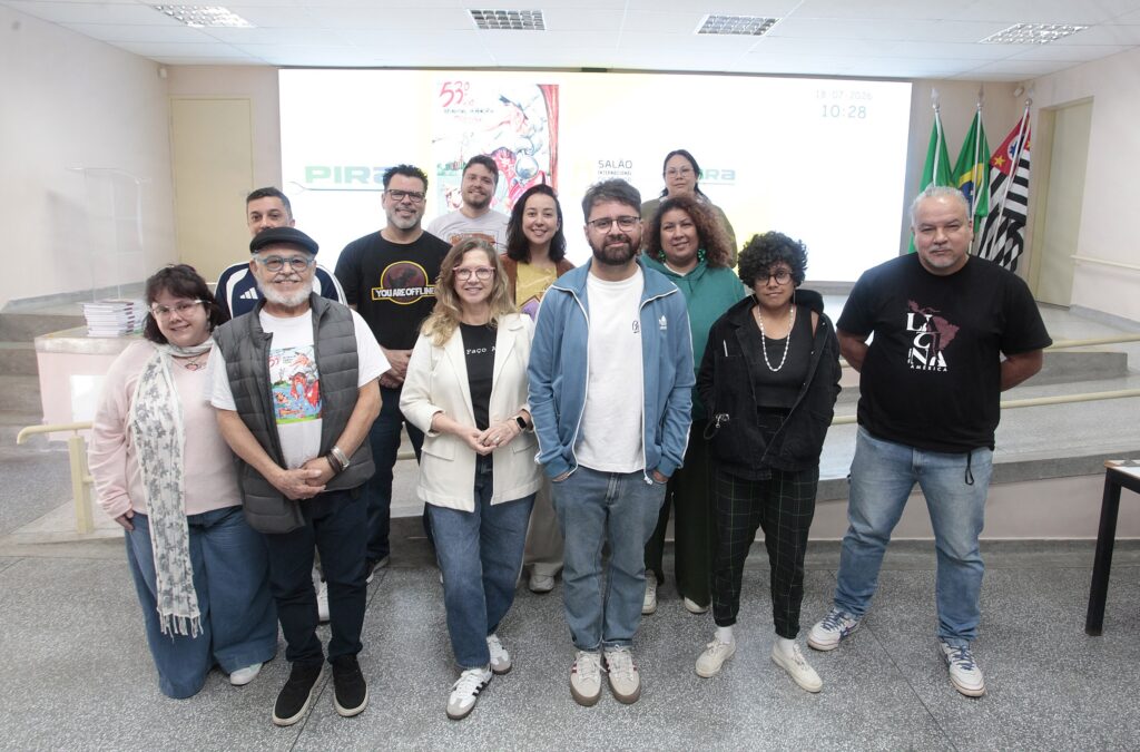 Esta foto mostra um grupo de 13 pessoas posando em um ambiente interno, tipo auditório ou sala de eventos, em frente a uma grande tela branca iluminada. Composição: três filas informais — quatro pessoas na frente, cinco na segunda fila e quatro atrás — todas voltadas para a câmera, com expressões majoritariamente sorridentes ou neutras. Ambiente: palco baixo com degraus ao fundo, tela de projeção/monitor grande iluminando a parede (há logotipos e, no canto direito superior, data e hora: "18-07-2026 10:28"). À direita, bandeiras (incluindo a do Brasil e de São Paulo) presas em mastros. Idades e diversidade: grupo variado em idade e aparência; há homens e mulheres, tons de pele diferentes e estilos diversos, sugerindo participação comunitária ou equipe multidisciplinar. Roupas e estilo: visual casual — camisetas, jaquetas esportivas, suéteres, colete, blazer claro e jeans. Alguns detalhes chamativos: óculos coloridos no homem de barba branca e boina, camiseta com estampa "You Are Offline" em um homem ao centro/esquerda, e uma mulher com brincos verdes grandes atrás à direita. Expressão e postura: postura descontraída e amistosa; mãos à frente de alguns, braços atrás de outros; ninguém faz gestos extremos — parece foto oficial de encerramento ou registro de reunião. Elementos adicionais: ao fundo à esquerda há um púlpito de acrílico e uma pilha de livros/cadernos; piso em granito e corrimão claro ao lado do palco. Impressão geral: registro coletivo de um evento, oficina ou reunião institucional — atmosfera profissional, porém acolhedora e informal.