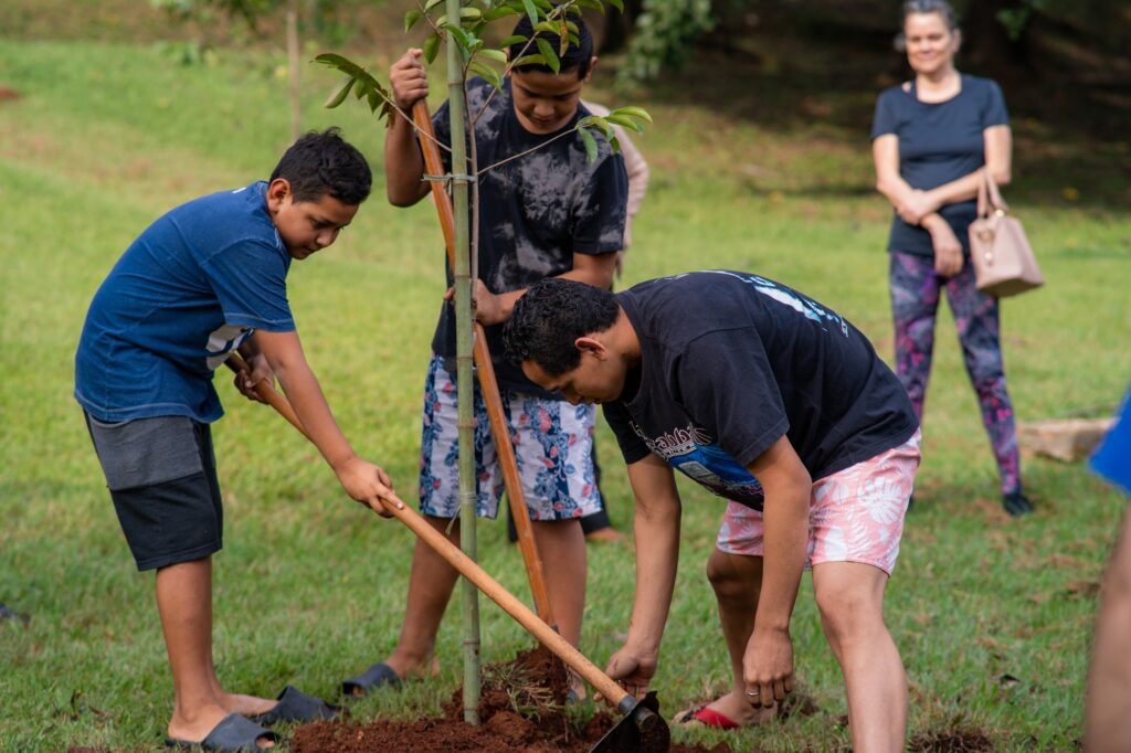 Esta é uma foto que registra um momento prático e colaborativo de educação ambiental. A Ação Principal: Três jovens estão trabalhando juntos para plantar uma árvore jovem. O rapaz à direita está agachado, usando uma enxada para trabalhar o solo ao redor da base da muda. O rapaz ao centro segura uma estaca de bambu que serve de suporte para a árvore, mantendo-a reta. O rapaz à esquerda ajuda com uma pá ou cabo de madeira, também auxiliando no processo de plantio. O Cenário: A atividade acontece em um gramado aberto e ensolarado. A muda de árvore, que parece ser nativa, está centralizada na cena. Observadora: Ao fundo, um pouco afastada, uma mulher está em pé, com os braços cruzados, observando a atividade com um sorriso tranquilo. Ela veste uma camiseta preta e calças estampadas, segurando uma bolsa. Atmosfera: A cena transmite cooperação, aprendizado e cuidado com a natureza. Os jovens estão focados na tarefa, mostrando um exemplo positivo de trabalho em equipe para o meio ambiente.