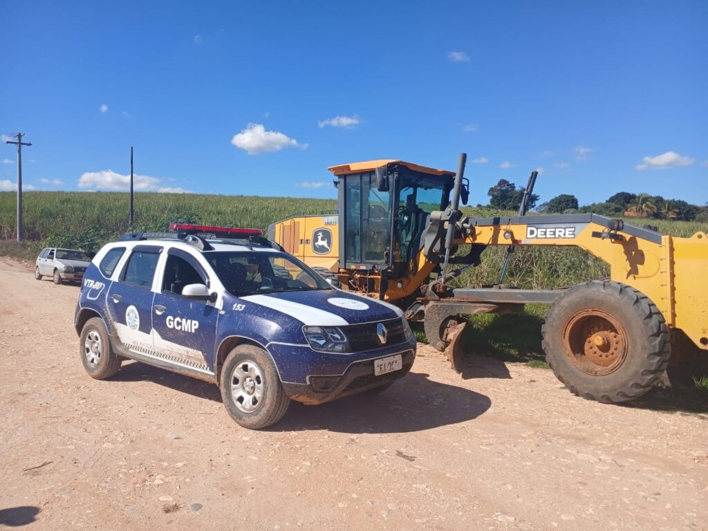 Foto de um trecho de estrada de terra em área rural com dois veículos em destaque: um carro oficial e uma motoniveladora. Principais pontos: Veículos: À esquerda, um SUV policial/veicular institucional em azul e branco com sirene no teto e identificação “GCMP” na lateral; mostra sujeira de estrada nas laterais e pneus. À direita, uma motoniveladora amarela marca John Deere (logo e a palavra “DEERE” visíveis) estacionada ao lado do SUV; pneus grandes com sujeira/oxidação. Ação: ambos estão parados sobre o cascalho/terra — parece uma vistoria, manutenção de via ou intervenção em estrada rural. Ambiente: campo agrícola ao fundo (provavelmente canavial ou plantação alta), postes de energia ao longo da estrada e vegetação mais densa ao longe. Céu e clima: céu azul limpo com poucas nuvens pequenas; luz do dia forte e sombras bem definidas, indicando dia ensolarado. Detalhes visuais: estrada de superfície irregular com pedras soltas; um carro prata estacionado mais atrás; a motoniveladora apresenta marcas de uso; o SUV tem numeração “153” visível na lateral. Observação de privacidade: não há pessoas identificáveis na cena; não posso nomear indivíduos.