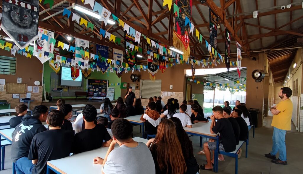Sala ampla tipo refeitório/auditório escolar, decorada com muitas bandeirolas e estandartes pendurados no teto, com mesas compridas e bancos onde um grande grupo de jovens está sentado, assistindo a uma apresentação. Disposição: várias fileiras de mesas brancas com estrutura azul ocupadas por alunos adolescentes vistos de costas e de perfil; todos voltados para a frente onde há um pequeno palco/área de apresentação. Apresentador: uma pessoa de uniforme escuro em pé à frente, falando perto de um projetor; não é possível identificá‑la nem afirmar sua função. Recursos visuais: projetor ligado exibindo slides numa tela ao fundo; um painel expositivo e quadros na parede próximos ao palco. Decoração e teto: estrutura de madeira aparente com vigas; bandeirolas coloridas (amarela, verde, azul, branca) e estandartes decorativos pendem por todo o salão, dando clima festivo ou de evento escolar/associação. Público e comportamento: alunos atentos, alguns apoiados nos braços, outros cochichando baixinho; há também um adulto de pé à direita (camiseta amarela) observando a apresentação com os braços cruzados. Iluminação e ambiente: luz natural entrando pelas aberturas laterais e iluminação interna; piso e paredes com aspecto funcional — ambiente escolar ou comunitário, descontraído e organizado.