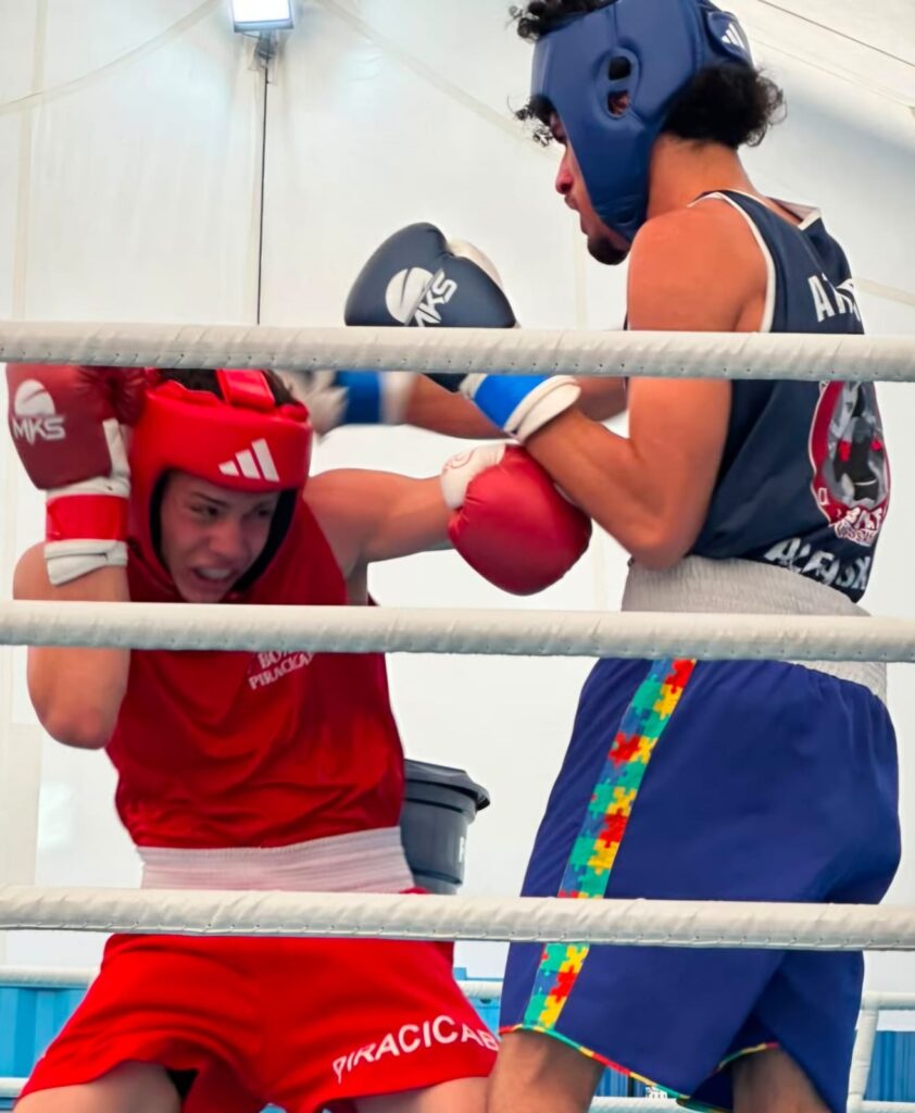 Foto de uma luta de boxe amadora dentro do ringue. Observações detalhadas: Dois pugilistas de frente, um de vermelho (à esquerda) e outro de azul (à direita). Lutador de vermelho ajoelhado/agachado, protegendo a cabeça com a luva esquerda e a outra apoiada na lateral do rosto; expressão tensa e concentrada. Lutador de azul em posição ofensiva, braço direito avançado e luva direita quase tocando o oponente; usa protetor de cabeça azul e camiseta/regata azul escura. Ambos usam protetores de cabeça acolchoados e luvas de boxe visíveis (vermelhas e azuis), sinal de combate amador/regulamentado. Cordas do ringue cruzando a imagem em primeiro plano, dando sensação de proximidade e enquadramento dentro do ringue. Fundo claro (tenda ou estrutura iluminada), iluminação uniforme e sem sombras duras; foco na ação dos atletas. Detalhes de roupa: o short do lutador azul tem uma faixa lateral colorida com padrão tipo peças de quebra-cabeça; o short do lutador vermelho tem texto branco na perna. Movimento e tensão: imagem captura um momento de ataque/defesa, com continuidade de movimento sugerida pelo posicionamento corporal e expressão facial.