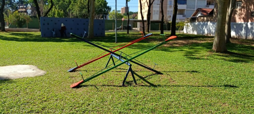 Esta imagem mostra um parquinho ao ar livre, com destaque para dois gangorras cruzadas em primeiro plano. Aqui estão os detalhes: As Gangorras: Estão posicionadas de forma a se cruzarem, formando um "X". Uma é verde com assentos vermelhos e a outra é preta com assentos vermelhos. Elas parecem ser feitas de metal e estão instaladas diretamente sobre o gramado. O Cenário: O local é um parque ou praça bem cuidado, com muita grama verde e árvores que projetam sombras longas, indicando que o sol está um pouco inclinado — talvez final da manhã ou tarde. Fundo: Ao fundo, há um muro baixo cinza, que parece ser uma parede de escalada com algumas pequenas aberturas, e algumas árvores. Mais atrás, é possível ver construções, o que sugere que o parquinho fica em uma área urbana, possivelmente residencial. Atmosfera: O dia está ensolarado e parece bastante tranquilo, convidando para uma brincadeira.