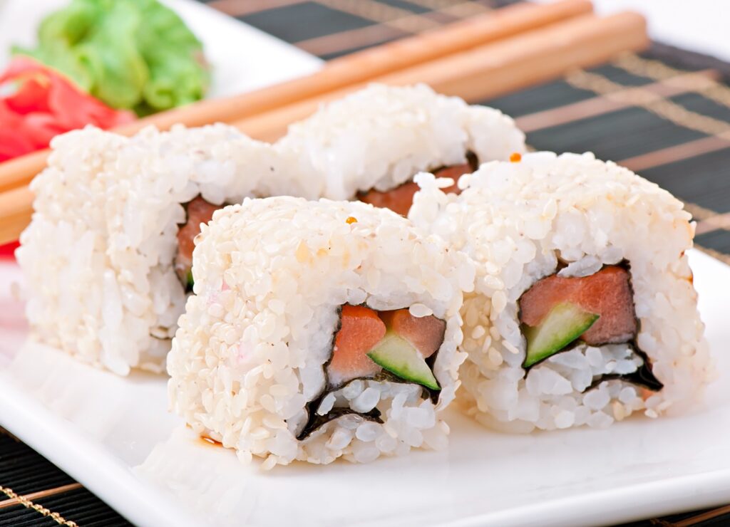 close-up de uramakis servidos em prato branco — rolinhos de sushi com arroz por fora e gergelim, recheio visível de peixe (possivelmente salmão) e pedaços verdes (abacate ou pepino). Pontos principais: Tipo: sushi do estilo uramaki (arroz do lado de fora, nori por dentro). Aparência: arroz coberto por sementes de gergelim; cortes generosos, textura úmida/brilhante. Recheio: fatias de peixe rosado e pedaços verdes (provável abacate ou pepino). Composição: palitos de madeira ao fundo e guarnições desfocadas (gengibre em conserva e folhas verdes). Atmosfera: foto gastronômica, apetitosa e bem iluminada, foco na textura do arroz e no recheio.