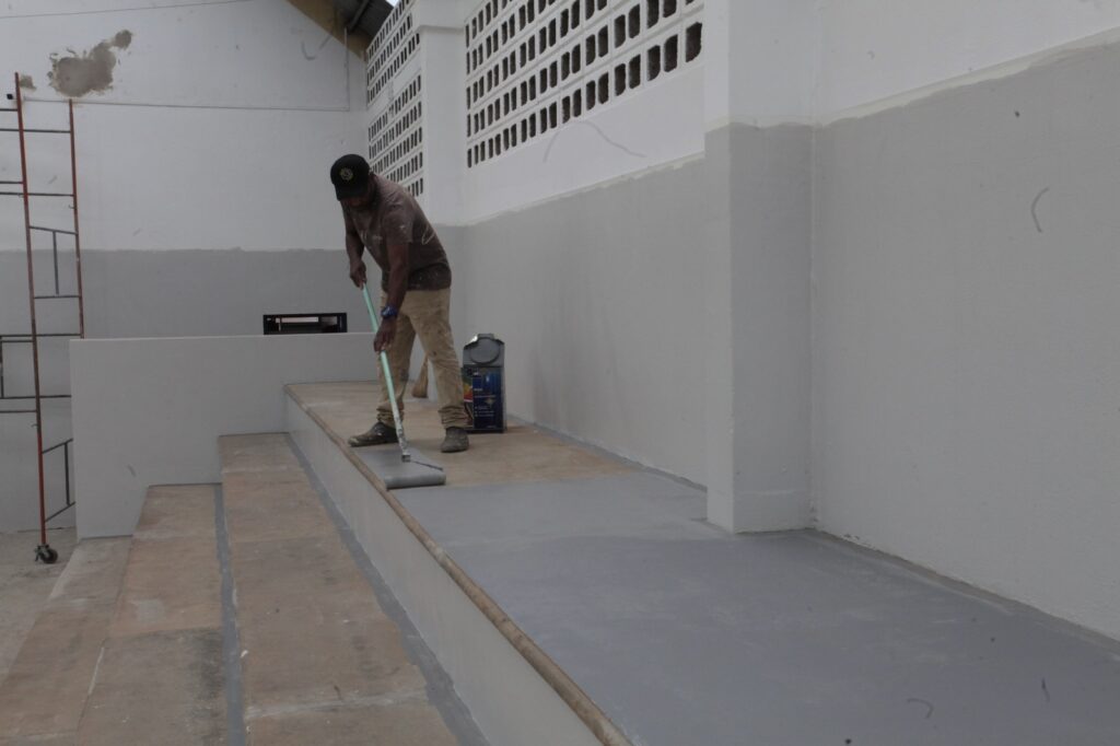 A imagem mostra um trabalhador pintando um patamar ou degrau elevado, provavelmente parte de uma arquibancada. Ele está usando um rolo de pintura com extensor para aplicar uma tinta cinza sobre a superfície. Ao lado dele, no chão, há uma lata de tinta aberta. O ambiente tem paredes pintadas em tons de cinza claro e branco, com uma estrutura de blocos vazados na parte superior da parede. À esquerda, vê-se parte de um andaime metálico. O espaço parece estar em reforma ou manutenção.