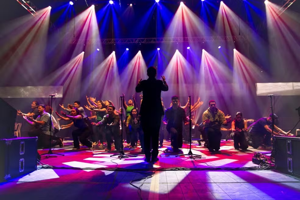 A imagem mostra um grupo de pessoas se apresentando em um palco, durante um show ou espetáculo.Detalhes da cena:Palco e iluminação: O chão é iluminado com luzes fortes em tons de vermelho, azul e branco. Vários refletores de cima criam feixes de luz que cortam a fumaça no ar, dando um efeito dramático. Tem caixas de som grandes nas laterais e microfones de pedestal espalhados.Apresentação: No centro, de costas para a câmera, está uma pessoa regendo ou dirigindo o grupo. Ela está de silhueta contra as luzes, com os braços levantados.Grupo: Ao redor do regente, cerca de 15 pessoas estão em formação. Algumas estão ajoelhadas, outras em pé com os braços abertos ou levantados. Elas usam roupas casuais e coloridas e parecem estar cantando, dançando ou fazendo uma performance teatral/coral. A expressão é de energia e envolvimento.Atmosfera: O clima é de apresentação ao vivo, com movimento e emoção. As luzes e as poses passam sensação de força e união do grupo.Parece ser um coral, grupo de teatro musical ou apresentação de dança contemporânea em um teatro ou evento.