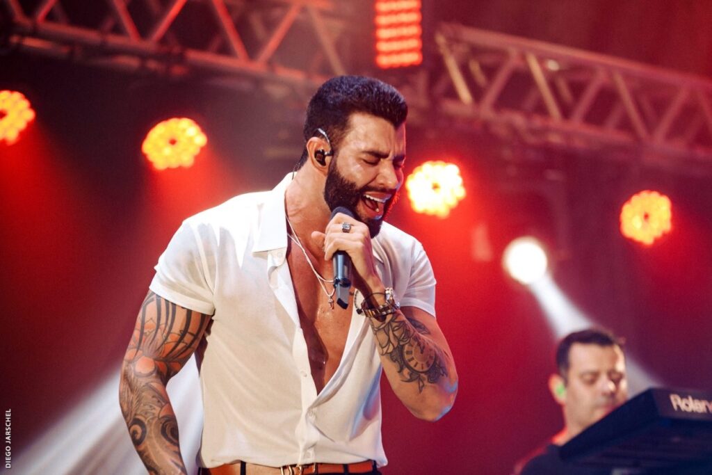 Esta imagem captura um momento intenso de uma apresentação ao vivo do cantor sertanejo Gusttavo Lima. Aqui estão os detalhes: O cantor: Gusttavo Lima aparece no centro, com os olhos fechados e cantando com emoção em um microfone. Ele usa uma camisa branca de botões (parcialmente aberta), revelando tatuagens elaboradas em ambos os braços, um colar com um pingente de cruz e um relógio robusto no pulso. O cenário: Ele está em um palco com iluminação cênica forte em tons avermelhados e estruturas metálicas (treliças) visíveis acima. Ao fundo, à direita, é possível ver parcialmente um músico tocando teclado (com a marca "Roland" visível). Créditos: No canto inferior esquerdo, há a assinatura do fotógrafo: "DIEGO JARSCHEL".