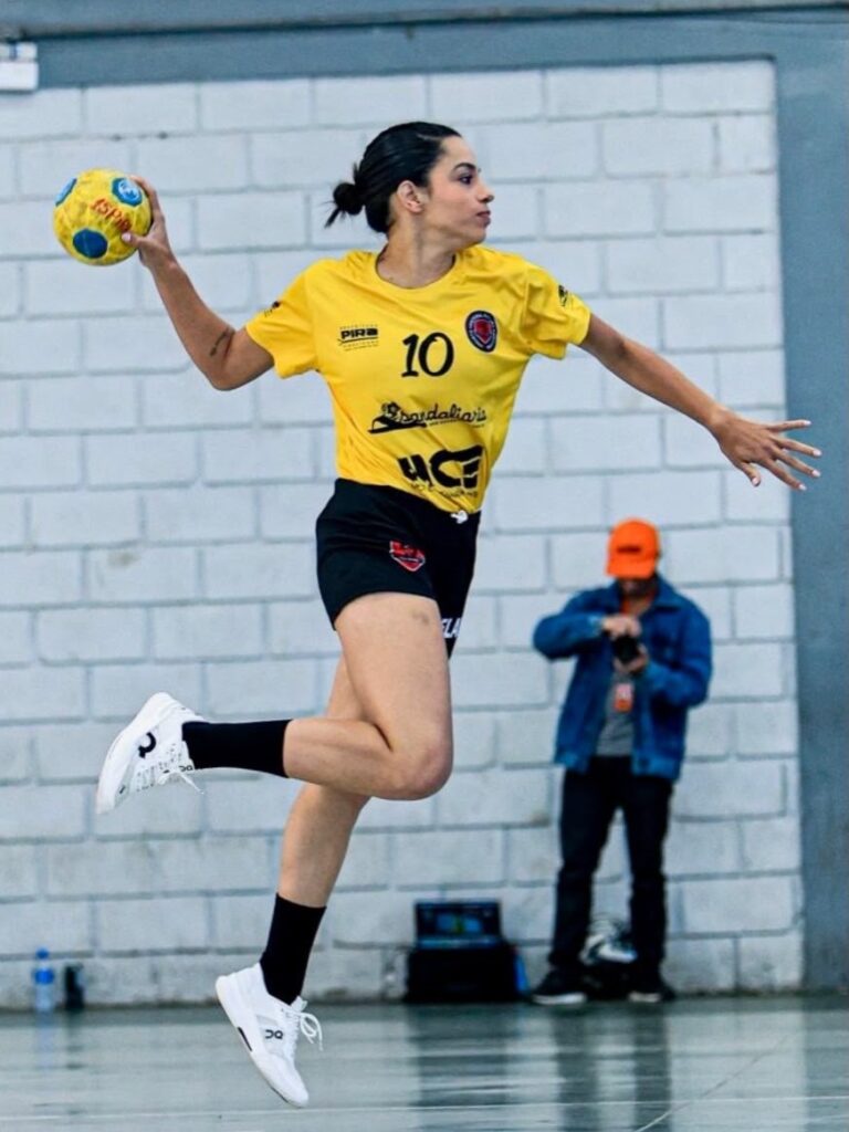 Uma jogadora de handebol em pleno movimento, capturada no ar durante um arremesso. A jogadora: Ela veste uma camisa amarela com o número "10" e um short preto. Seu cabelo está preso e ela está focada, com o braço armado para lançar uma bola de handebol amarela e azul. Ela usa tênis brancos com detalhes pretos e meias pretas. O cenário: A cena acontece em uma quadra coberta com parede de tijolos brancos ao fundo. Detalhes adicionais: Ao fundo, um homem de boné laranja e jaqueta jeans observa a cena, segurando uma câmera fotográfica, sugerindo que é uma cobertura esportiva profissional ou amadora. É uma foto que transmite muita energia e foco no esporte!