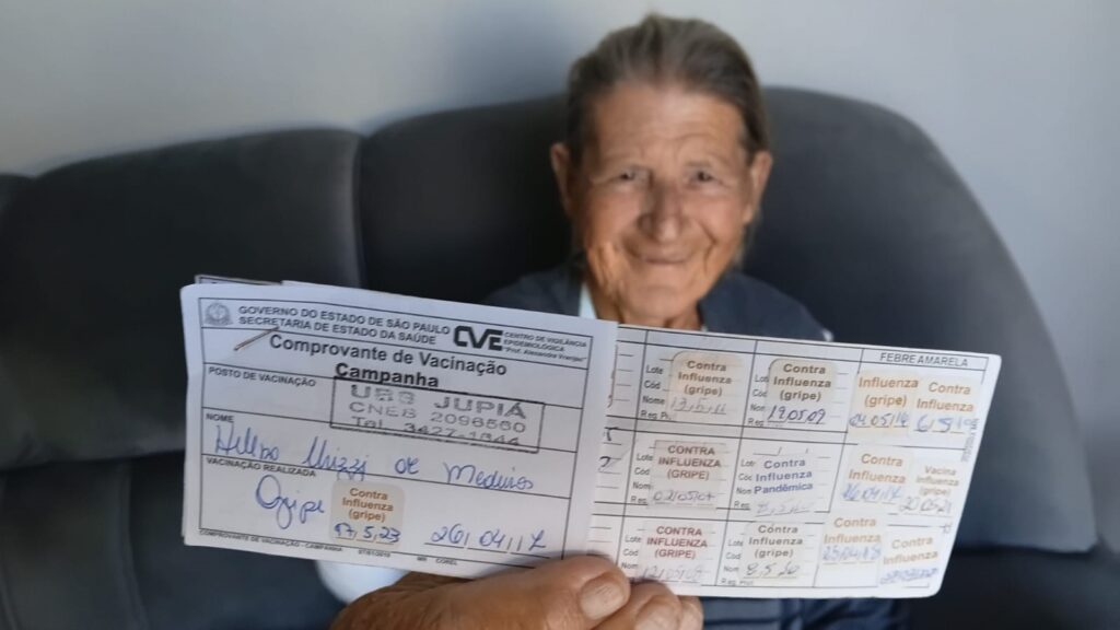 Visão geral: mulher idosa sentada numa poltrona/sofá de cor cinza escuro, sorrindo para a câmera e segurando um cartão de vacinação aberto em direção ao fotógrafo — o cartão está em foco no primeiro plano, o rosto dela aparece ligeiramente desfocado ao fundo. Pessoa: mulher adulta mais velha, cabelo preso para trás, pele enrugada típica da idade, expressão serena e acolhedora. Não posso identificar quem é. Cartão de vacinação (detalhes visíveis): Documento com o cabeçalho do Governo do Estado de São Paulo e menção à Secretaria de Estado da Saúde / CVE (Centro de Vigilância Epidemiológica). Título legível: "Comprovante de Vacinação — Campanha". Carimbo do posto de vacinação e espaços preenchidos com várias anotações manuscritas e carimbos. Diversos registros de doses contra influenza (gripe) preenchidos em vários quadros do cartão; também há referência a "FEBRE AMARELA" em uma seção. Várias datas e carimbos aparecem ao longo das células do cartão (algumas legíveis, outras parcialmente). O cartão parece registrar imunizações feitas em datas diferentes ao longo dos anos. Composição e foco: o cartão ocupa o primeiro plano, sendo segurado pela mão da mulher (dedos visíveis embaixo do documento); o recorte e o desfoque do fundo destacam o cartão como ponto focal. Atmosfera / tom: imagem transmite cuidado com a saúde, registro e memória de vacinação — sensação positiva e de orgulho ao mostrar o comprovante.