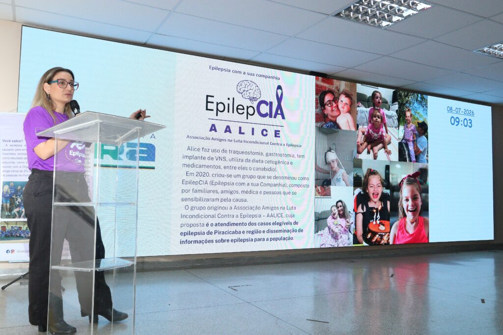 Cena interna de palestra/iniciativa de conscientização sobre epilepsia, com foco em uma palestrante em frente a um grande painel digital.
Pessoa/palestrante: mulher de cabelos longos claros, óculos e camiseta roxa segurando microfone com a mão direita; parece apontar/virar slides com um apontador na mão esquerda. Veste calça escura e está atrás de um púlpito acrílico transparente.
Púlpito: estrutura em acrílico com prateleiras visíveis; refletindo o piso polido.
Painel/slide: enorme tela LED ocupa quase toda a parede ao fundo. À esquerda do painel há um bloco de texto que exibe o título/logo "Epil epCIA" (EpilepCIA) e o nome "AALICE" — material informativo sobre epilepsia, com parágrafos explicativos. À direita aparecem várias fotos de crianças e famílias em formato de colagem, transmitindo histórias pessoais e rostos sorrindo.
Relógio/data: no canto direito da tela há data e hora exibidas — "08-07-2026" e "09:03".
Ambiente: piso liso e refletivo, teto com forro modular e luminárias embutidas; sensação de auditório ou sala de eventos institucional.
Atmosfera/propósito: aparência de evento público ou ação de sensibilização sobre epilepsia, combinando apresentação oral e materiais visuais com depoimentos/fotos de beneficiários ou participantes. Cena transmite profissionalismo e tom informativo.